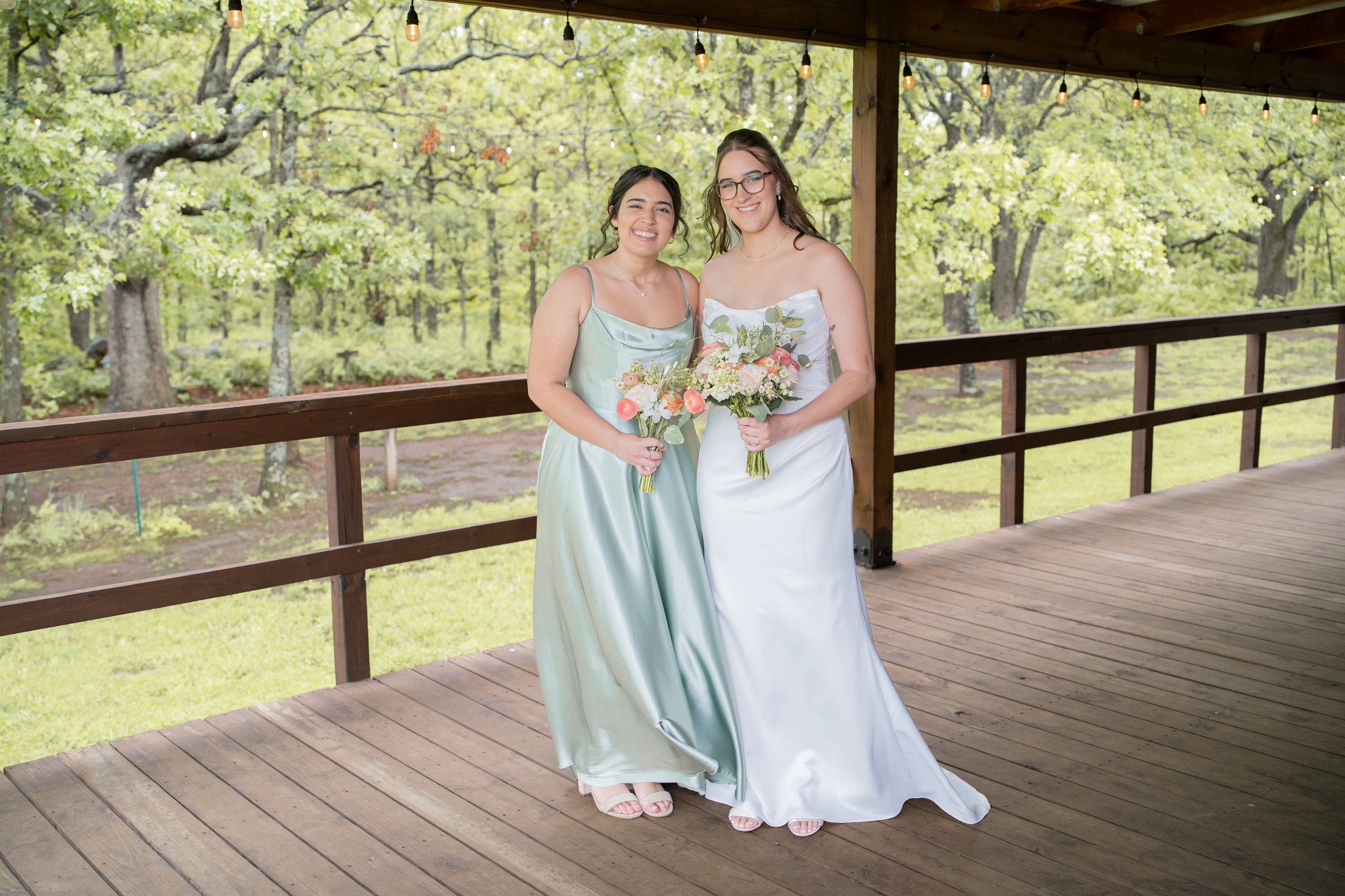 Two women in pastel-colored dresses holding bouquets, standing on a wooden porch with string lights, in a lush green outdoor setting.