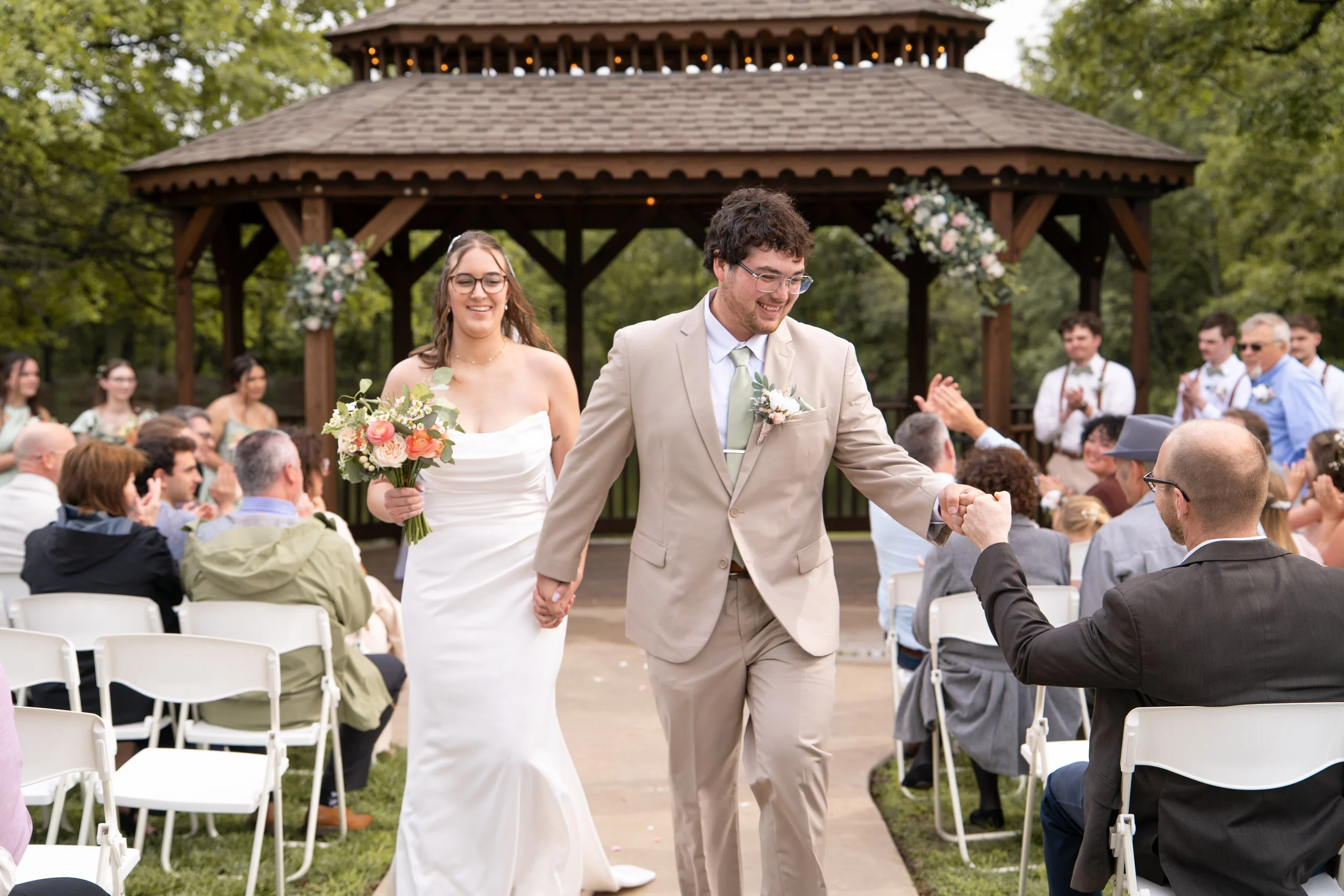 A newlywed couple walking hand in hand down the aisle of an outdoor wedding ceremony, smiling and holding hands with guests seated on either side, with a gazebo and greenery in the background.