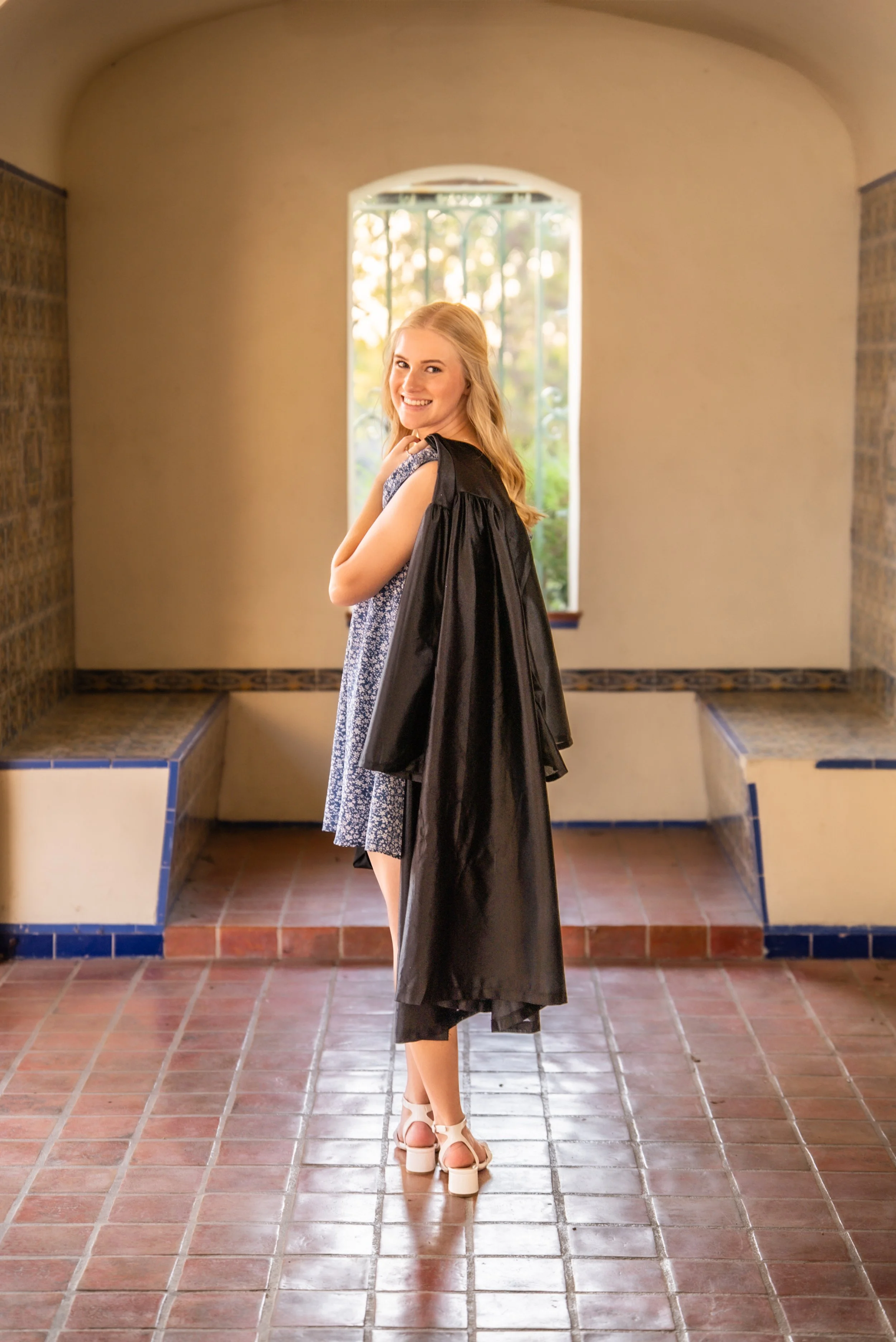 A young woman standing indoors, smiling, wearing a blue patterned dress and high heels, with a black graduation gown draped over her shoulders, in front of a window with a garden view.