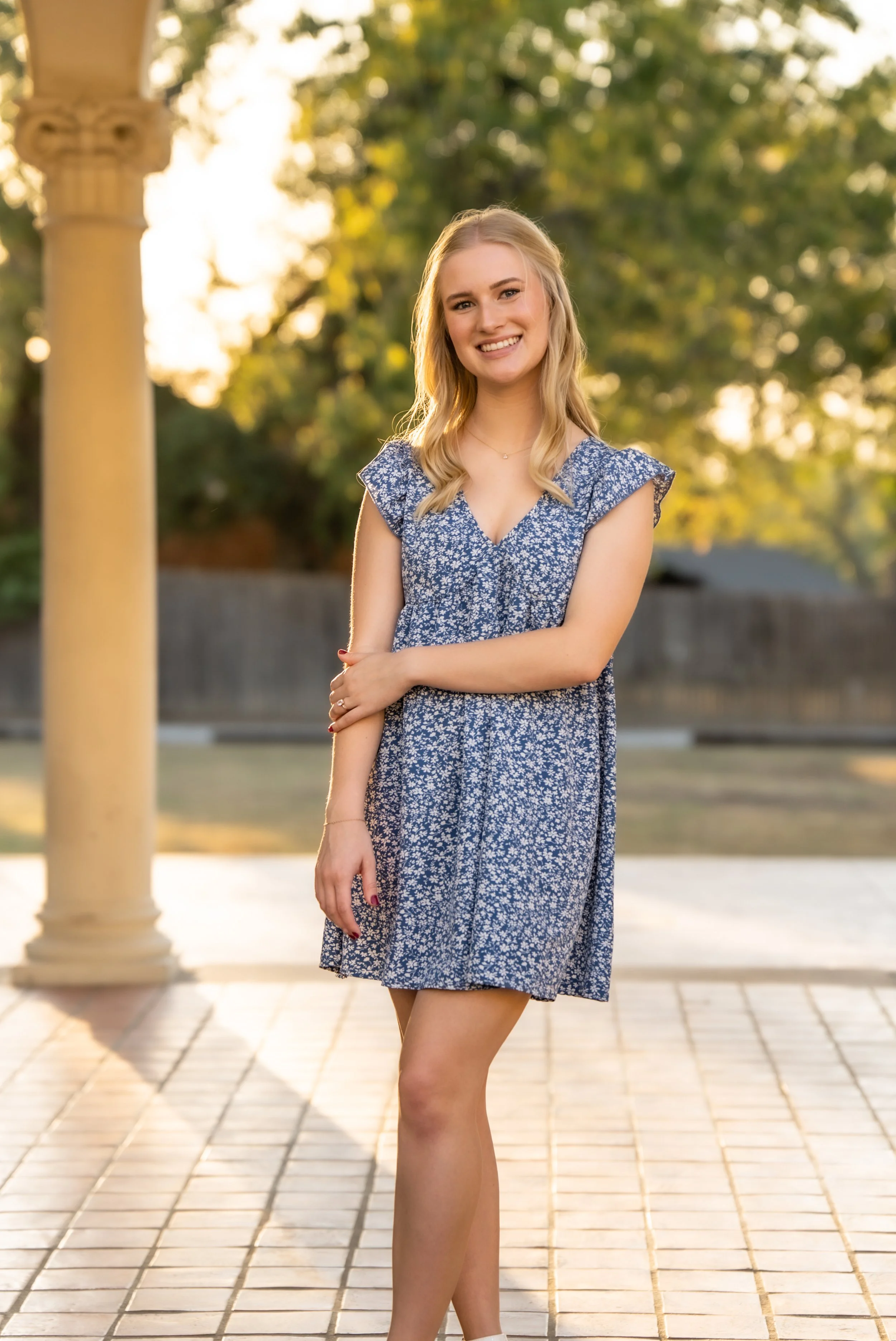 A young woman with blonde hair wearing a blue floral dress, smiling and standing outdoors on a paved area with trees and a column in the background during sunset.