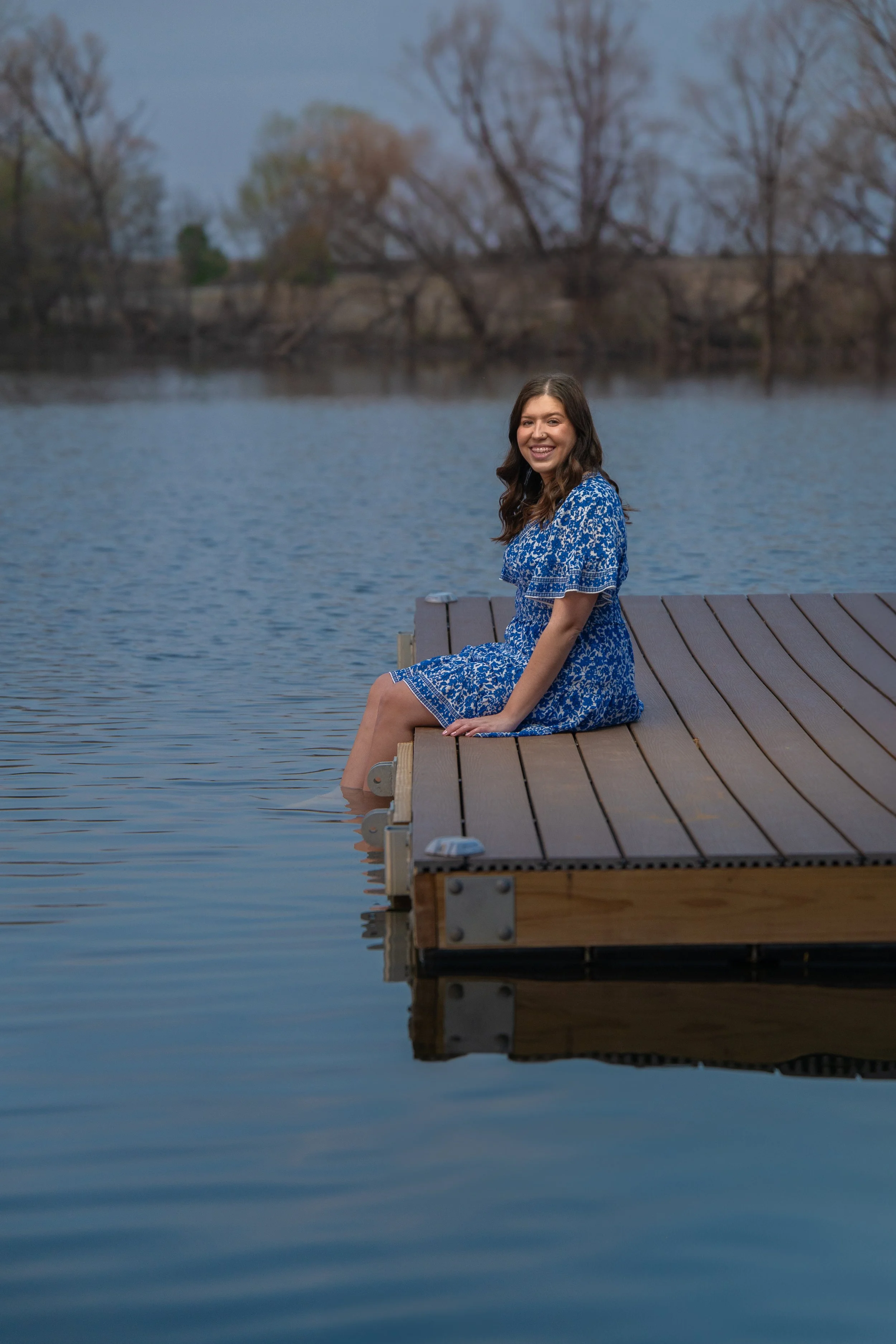 A young woman in a blue dress sitting on the edge of a wooden dock with her feet in the water, smiling, with a lake and trees in the background.