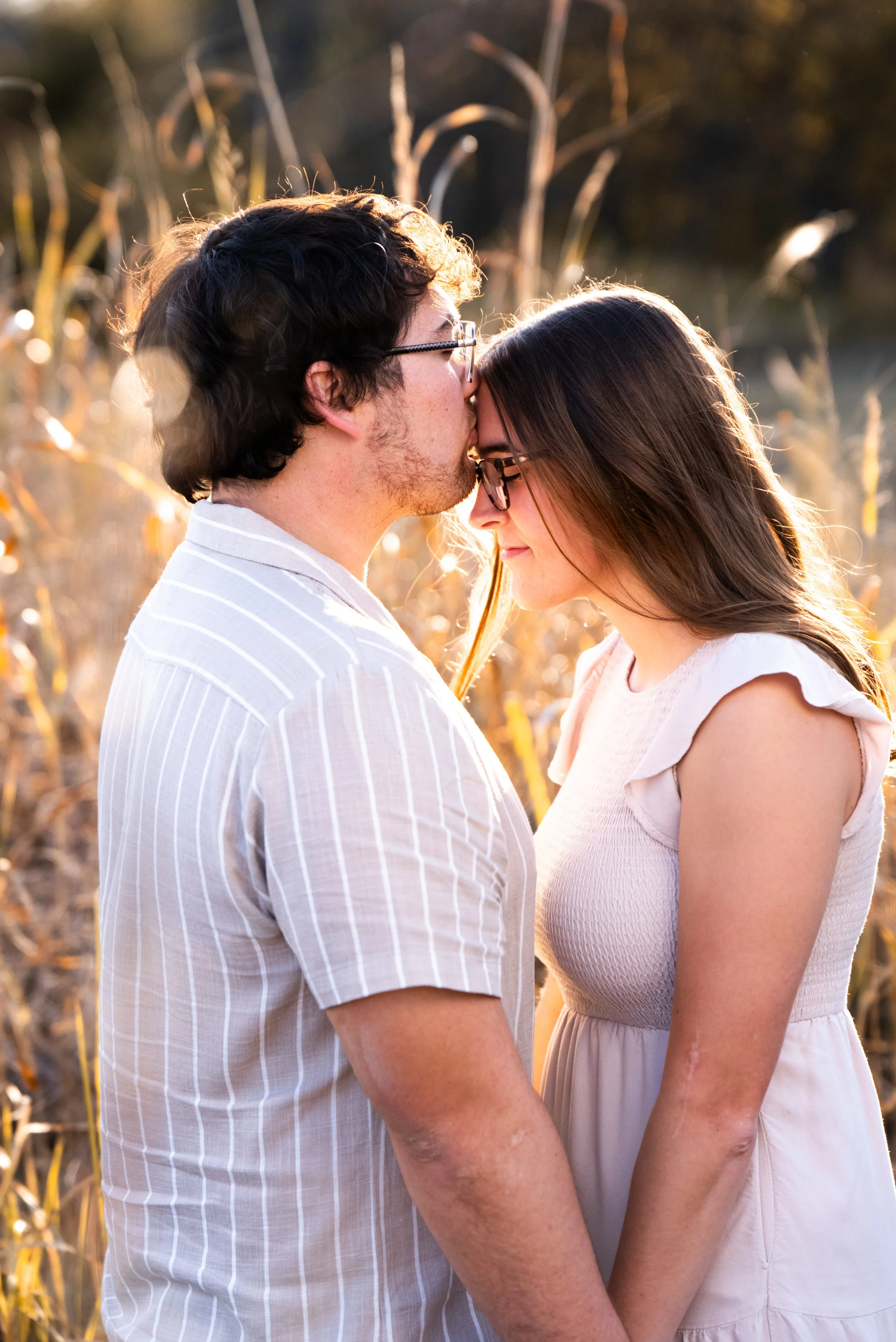 A couple standing close together in a field at sunset, with the man kissing the woman's forehead. Both are wearing glasses and light-colored clothing, and they are holding hands.