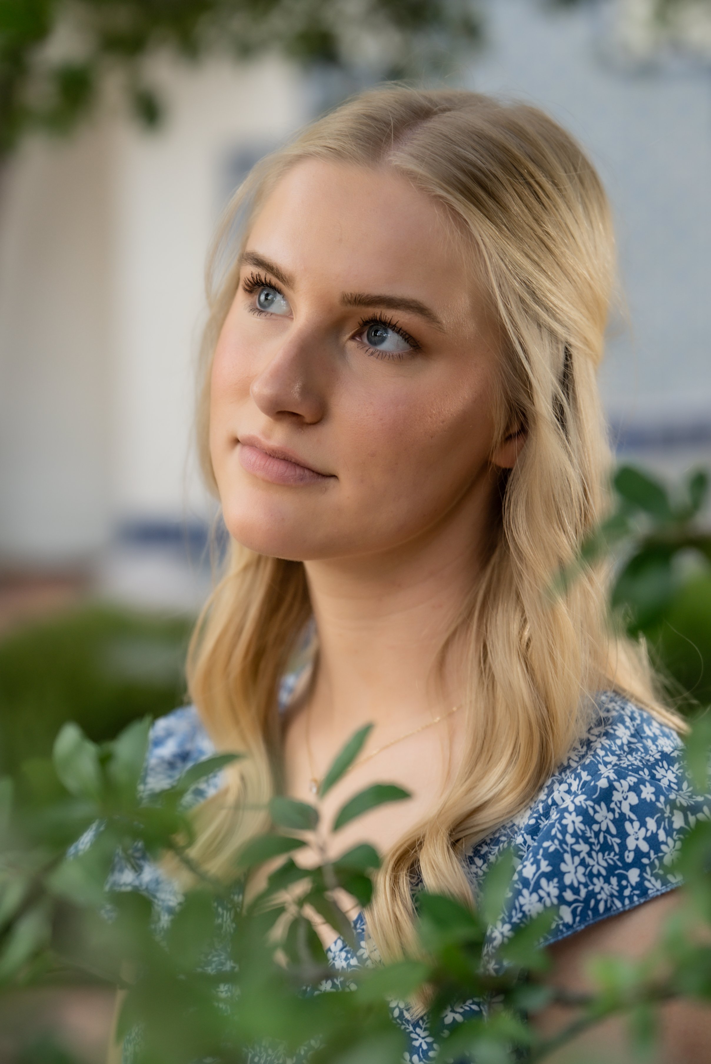 A young woman with blonde hair and blue eyes looking thoughtfully into the distance, surrounded by green foliage.