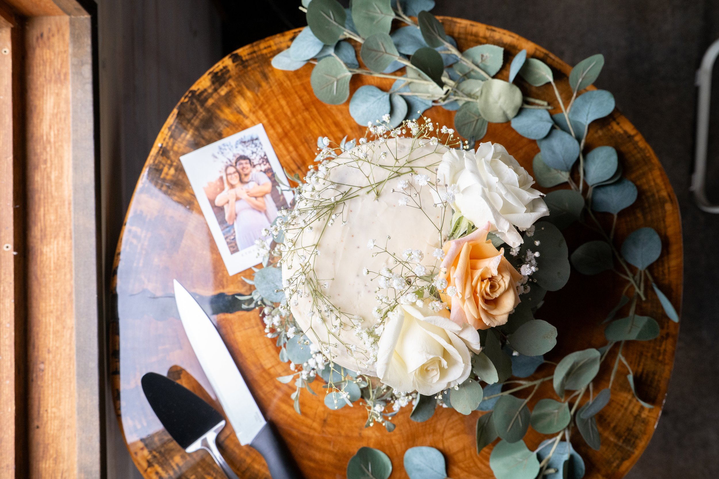 A wedding cake decorated with white, cream, and peach roses, baby's breath, and eucalyptus leaves on a wooden table, with a photograph and cake serving set nearby.