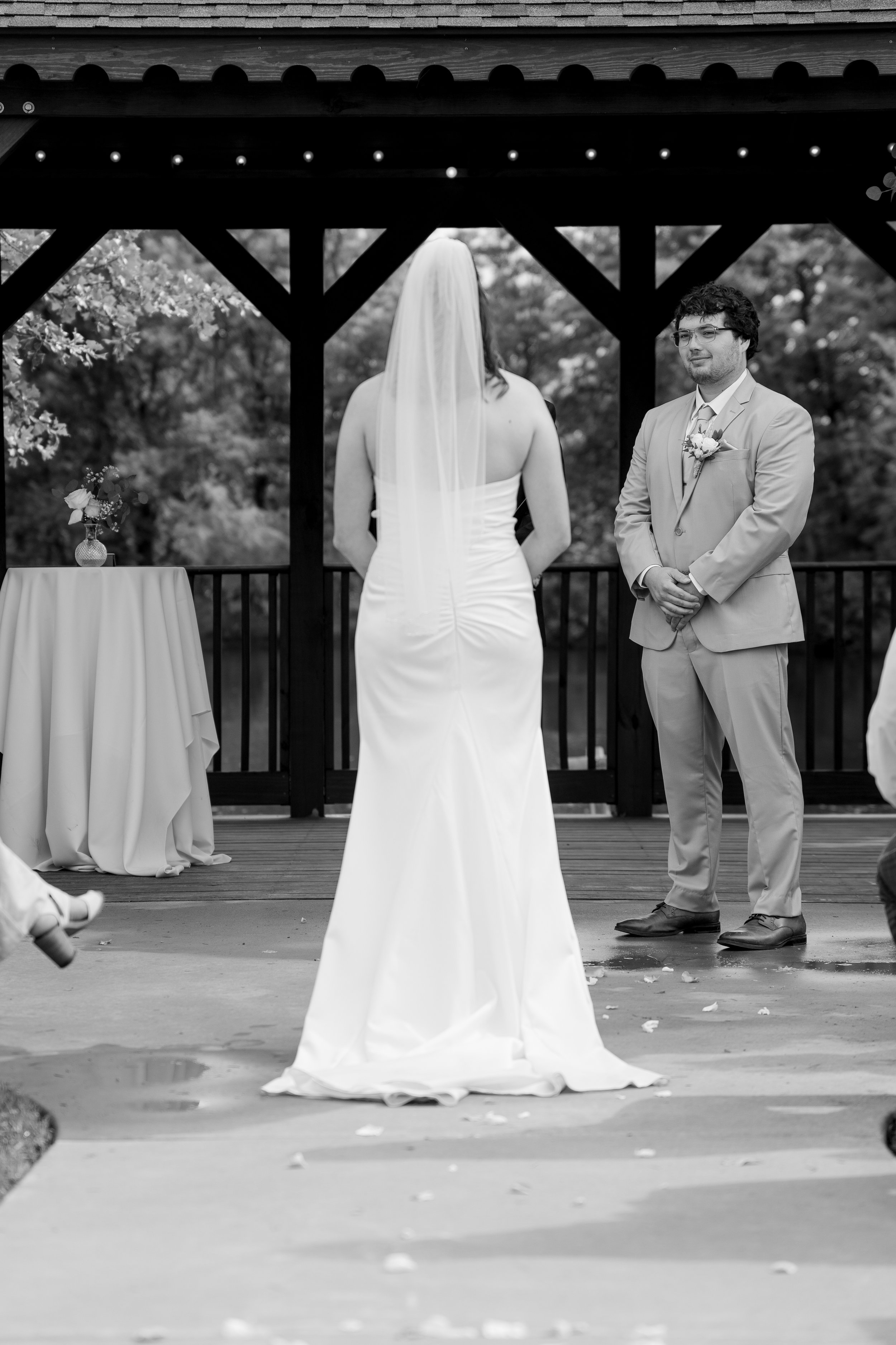 A black and white photo of a bride and groom during their wedding ceremony outdoors on a wooden deck, with trees in the background. The bride is wearing a strapless wedding dress with a veil covering her face, and the groom is dressed in a light-colo