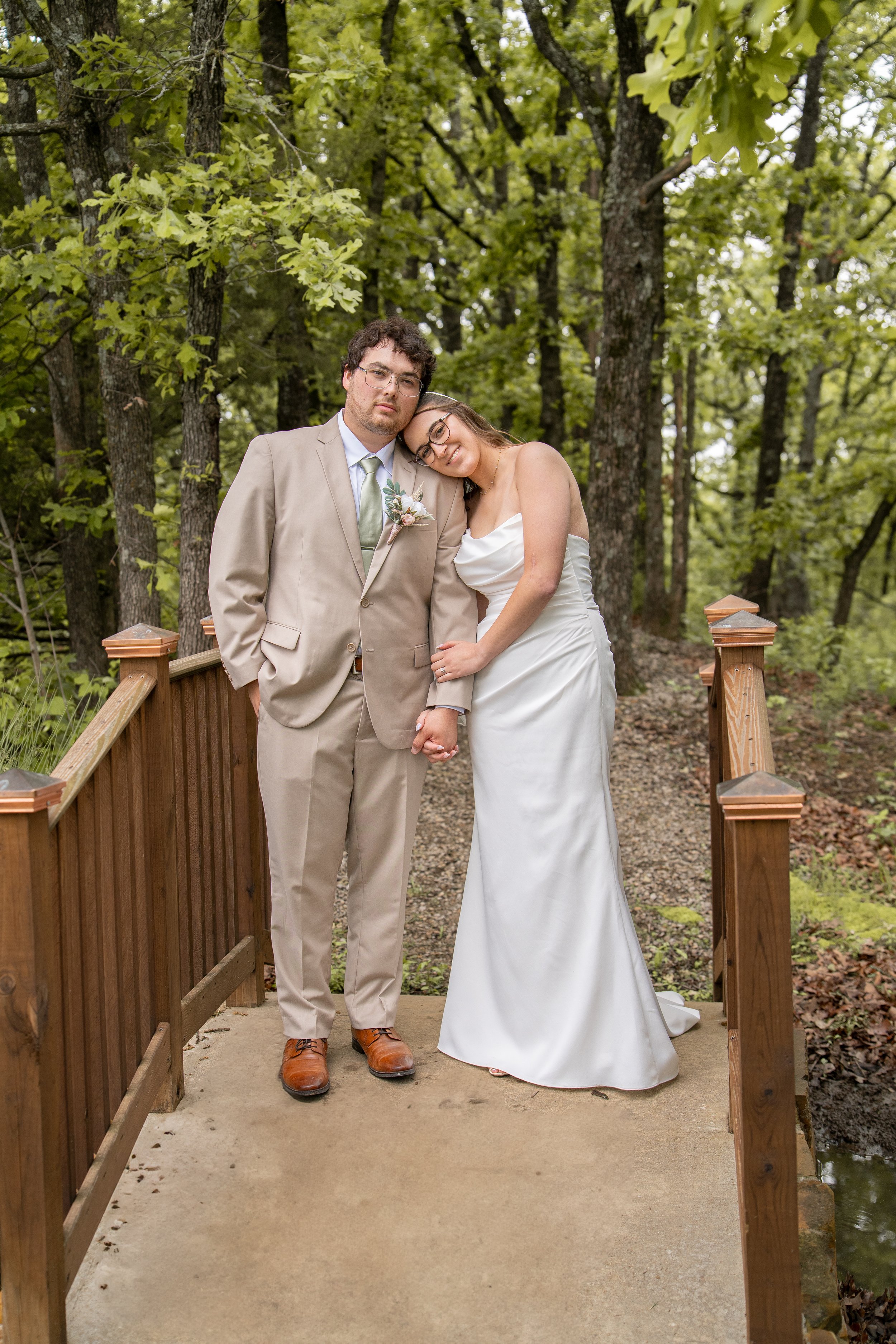 A couple standing on a wooden bridge in a forest during a wedding, the woman in a white wedding dress resting her head on the man's shoulder, holding hands.