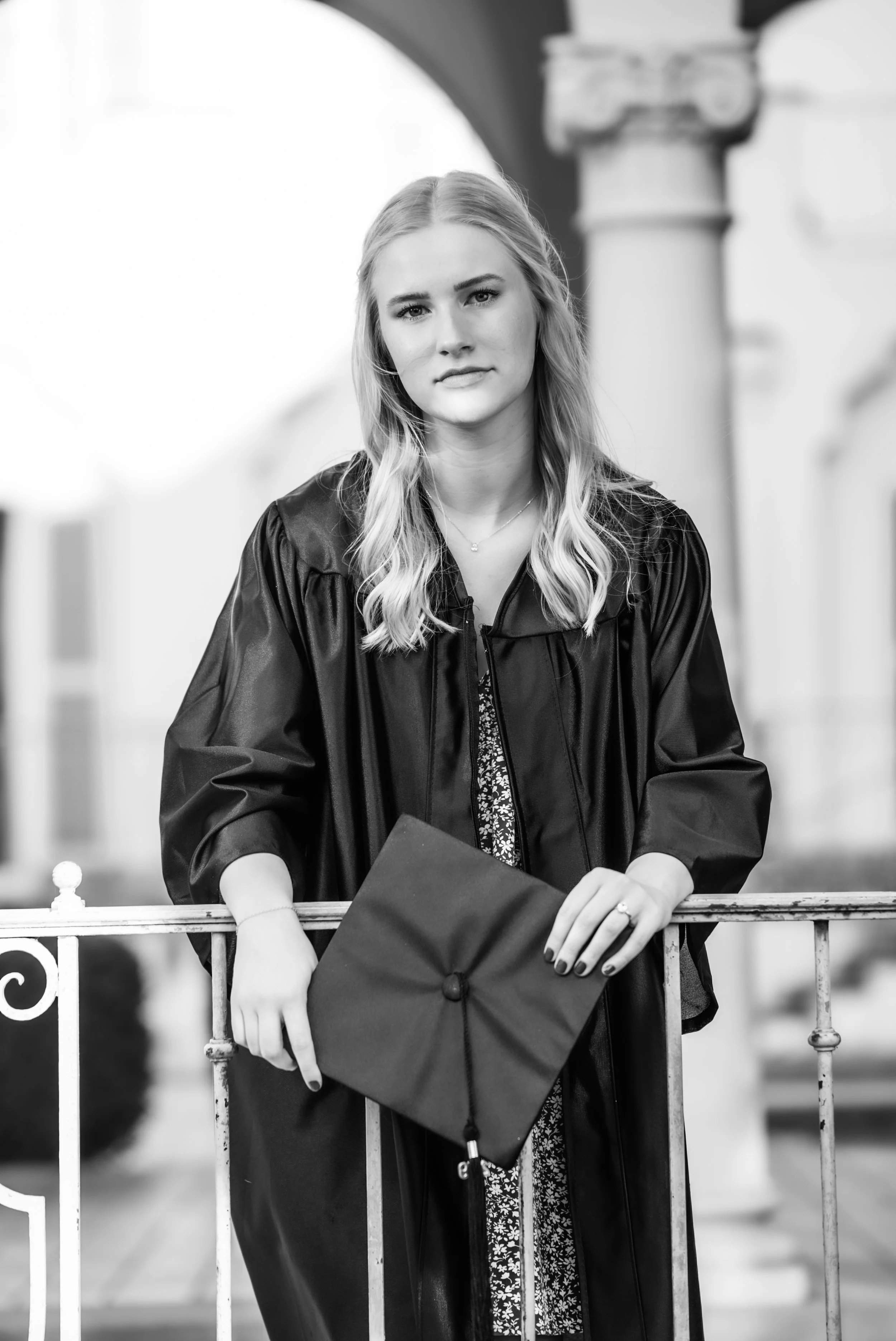 A young woman in a graduation gown holding a diploma or certificate, standing outdoors by a railing.
