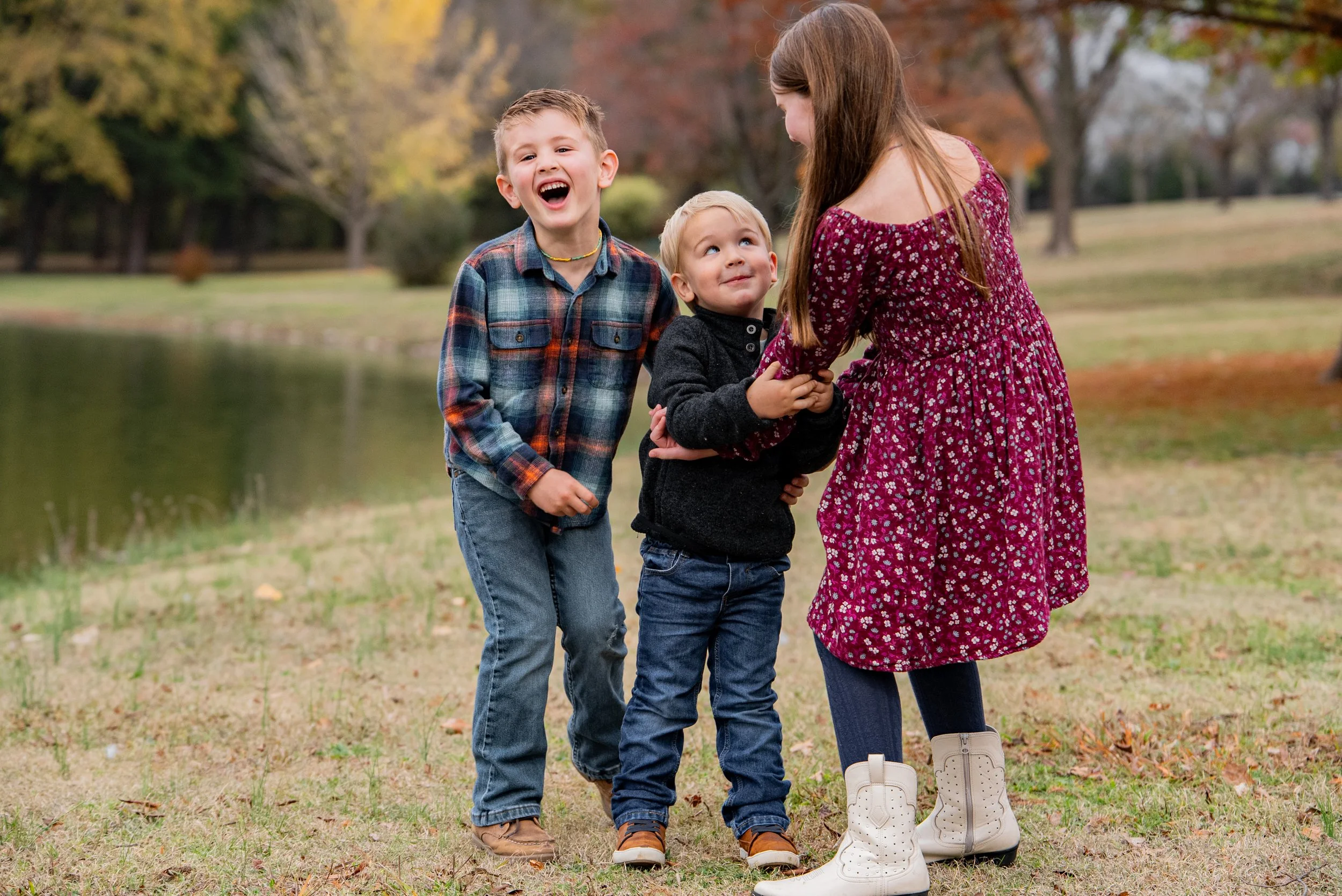 Three children laughing and playing together outdoors in a park with autumn trees and a pond in the background.