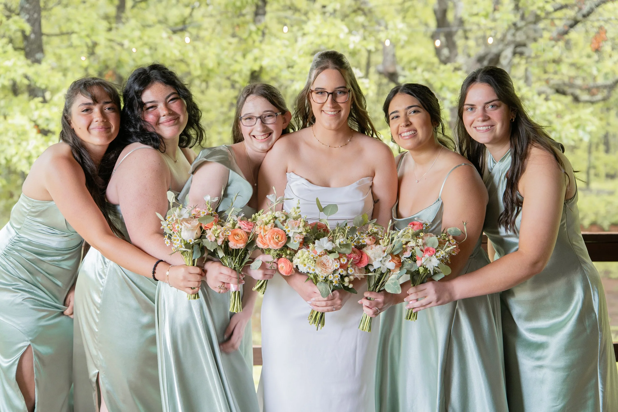 Group of six women in satin dresses holding bouquets of flowers, standing outdoors with green trees in the background.
