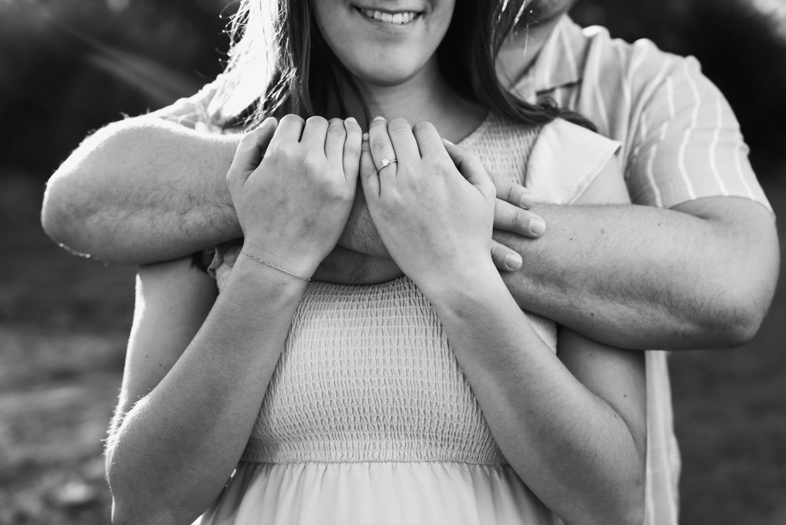 A woman with a smile receiving an embrace from a man, with her hands touching his arm, showcasing a wedding ring in a black and white photograph.