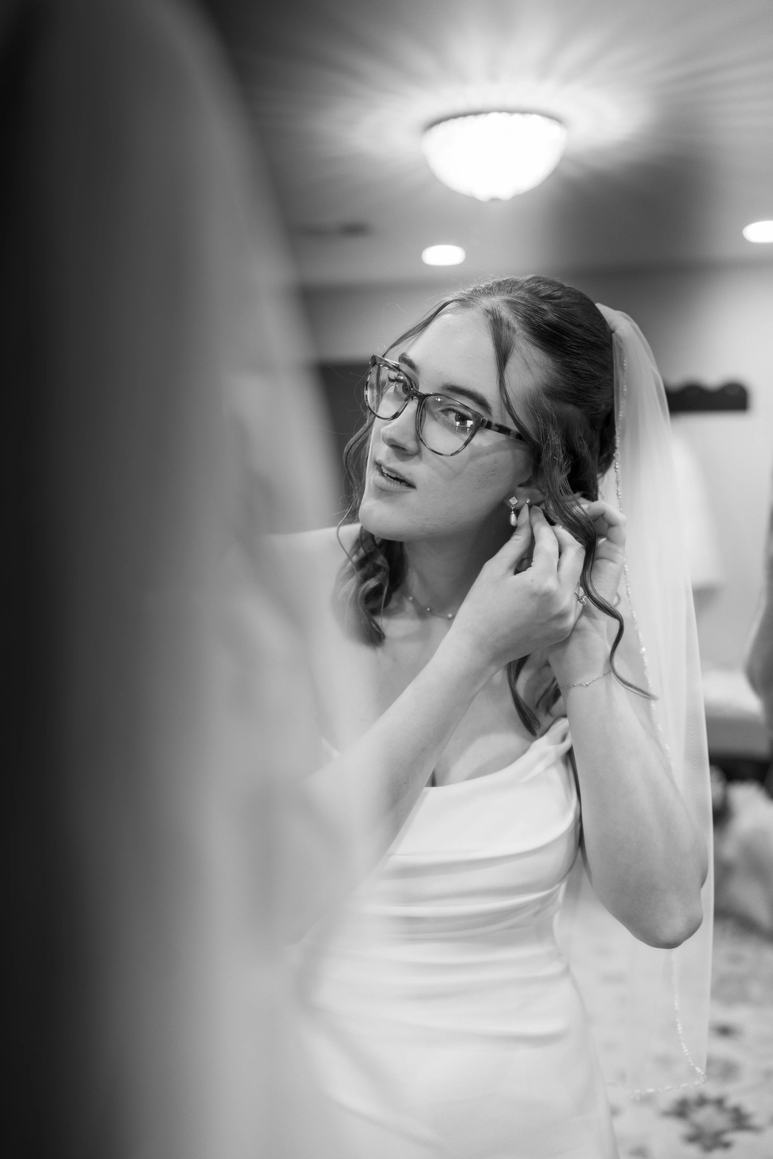 Black and white photo of a woman in a wedding dress putting on earrings, wearing glasses, and a veil, with a ceiling light in the background.