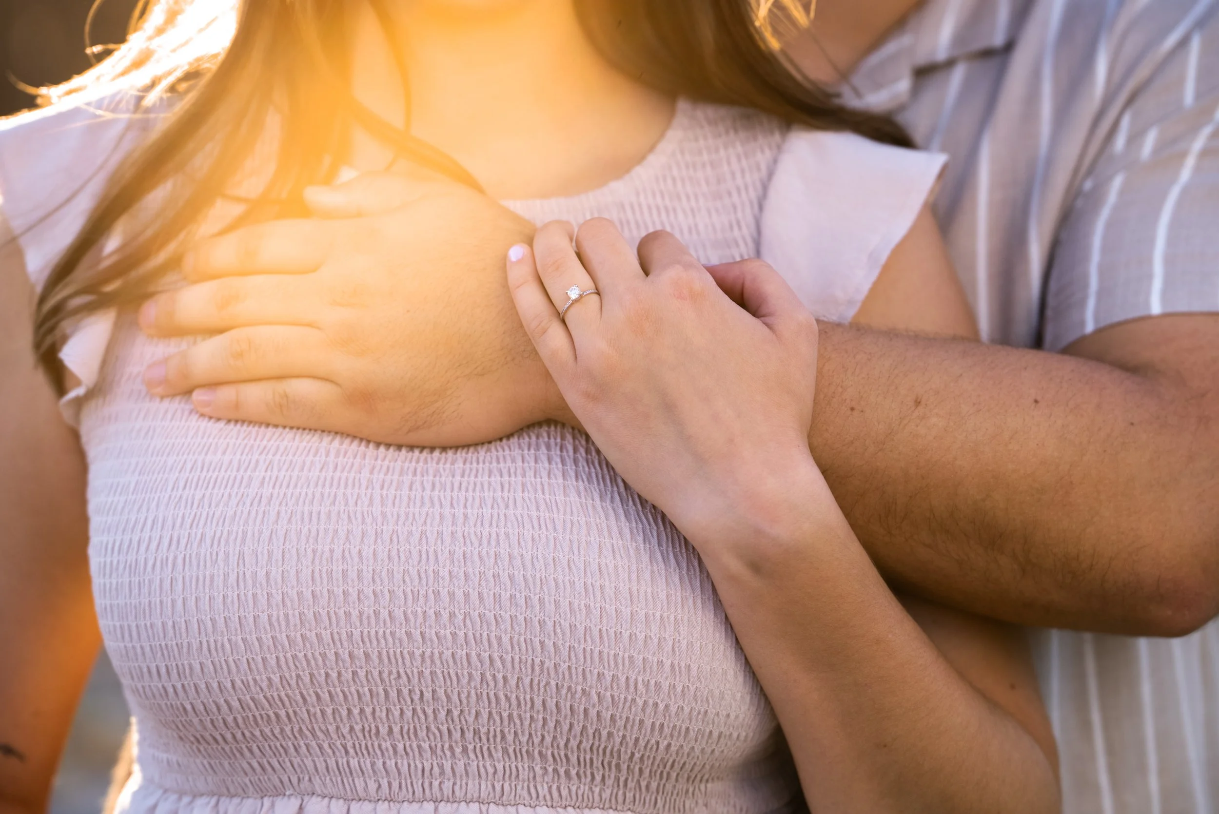 A woman with a ring on her finger places her hand on her chest while a man embraces her from behind.