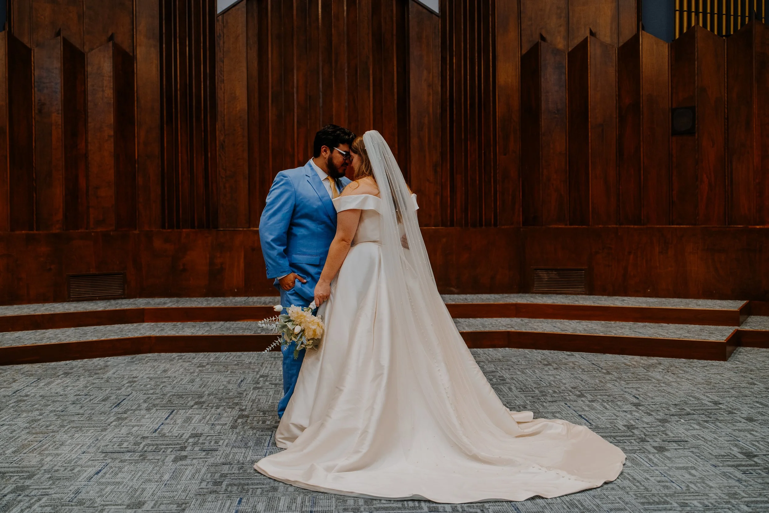 Bride and groom sharing a close, intimate moment in a wedding ceremony, standing on a carpeted floor with a wooden-paneled wall in the background. The bride is wearing a white off-the-shoulder gown with a long train and holding a bouquet, while the groom is in a bright blue suit.