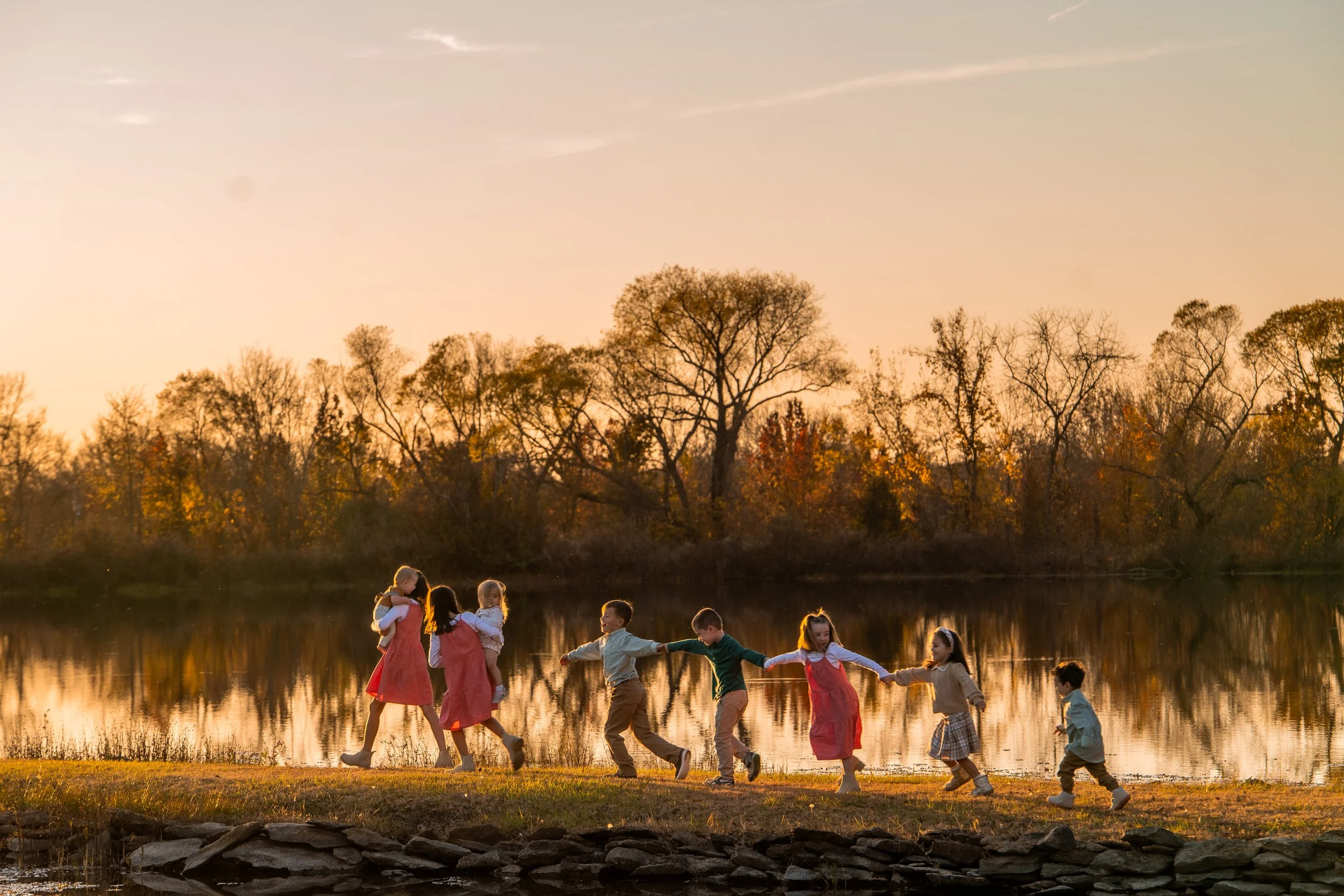 Children holding hands and walking along a lakeshore during sunset, with trees in the background.
