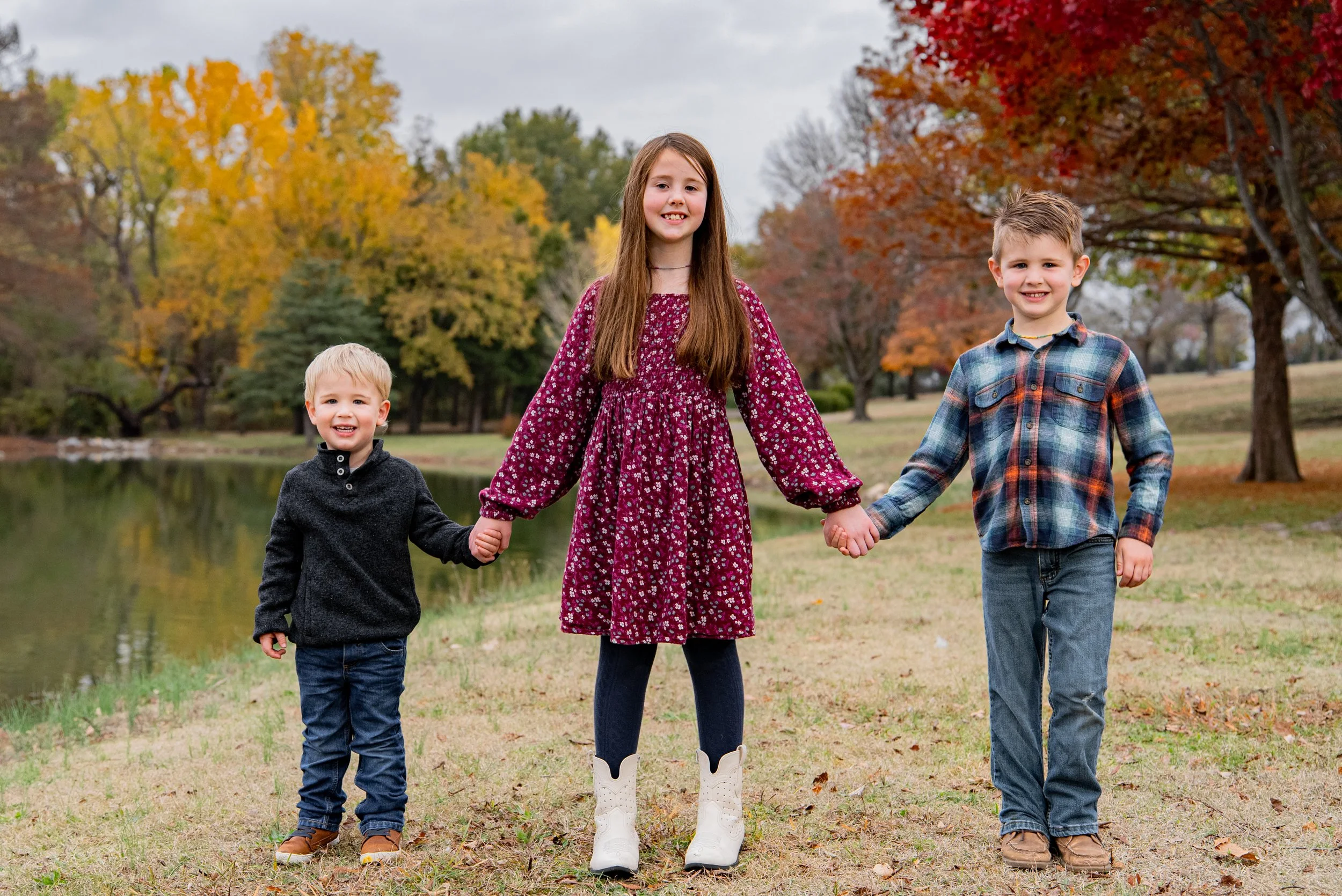 Three children holding hands and walking outdoors in a park during fall, with colorful autumn trees and a lake in the background.