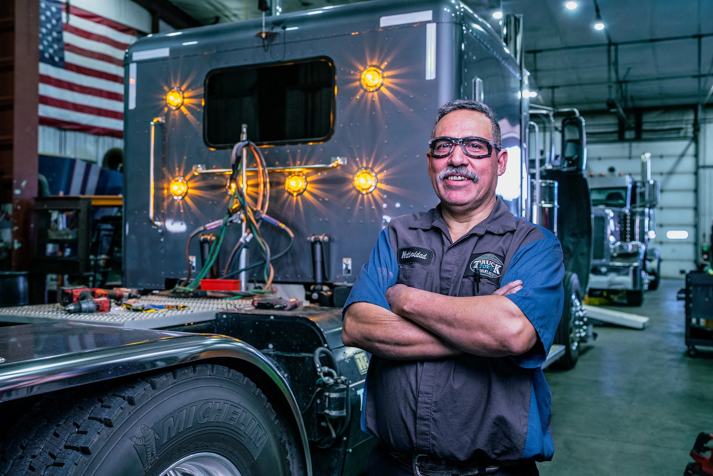 A man in work overalls standing in front of a large black semi-truck inside a warehouse, with American flag in the background.