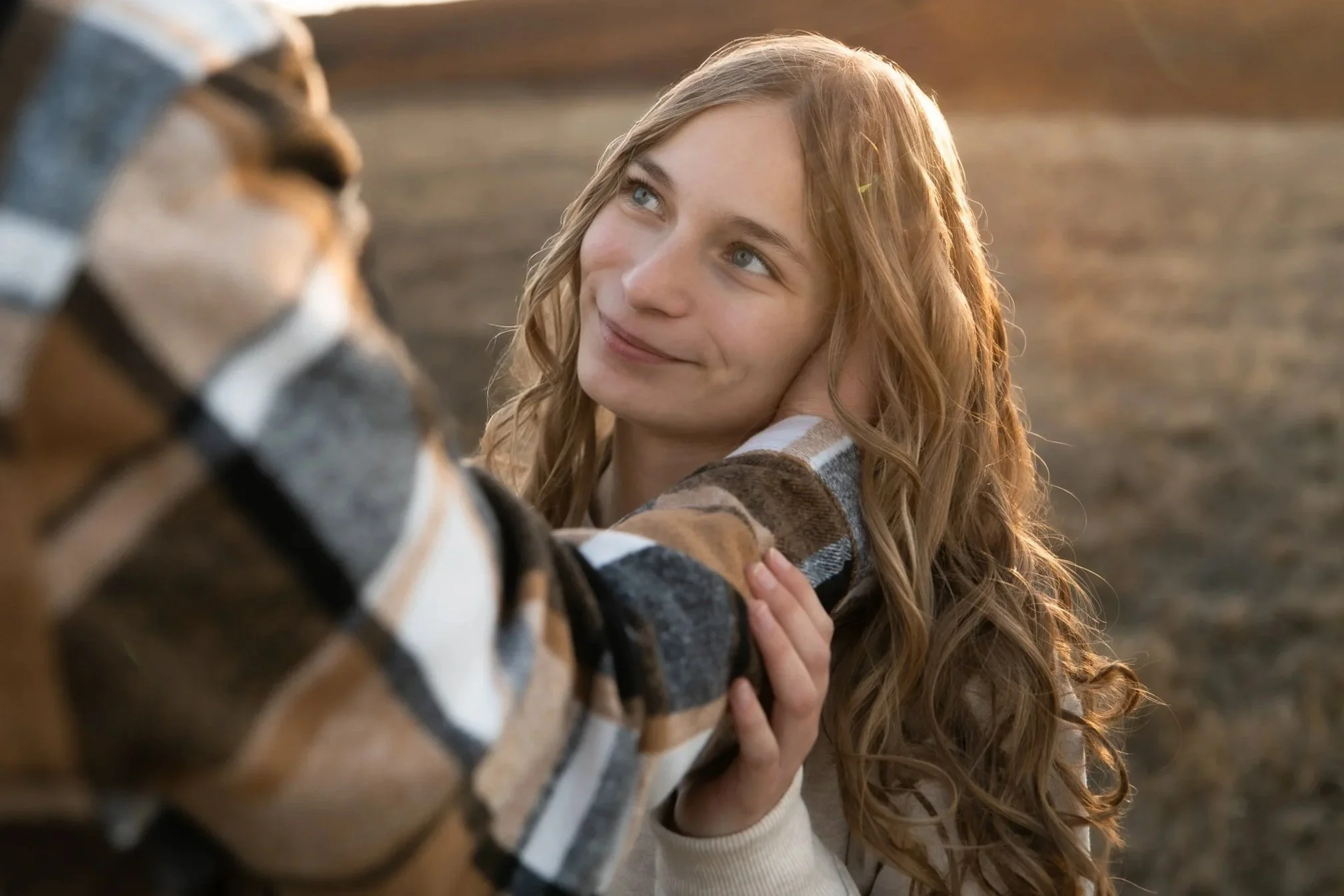 A young woman with long, curly red hair gazes affectionately at a dog, gently touching its face, outdoors during sunset.