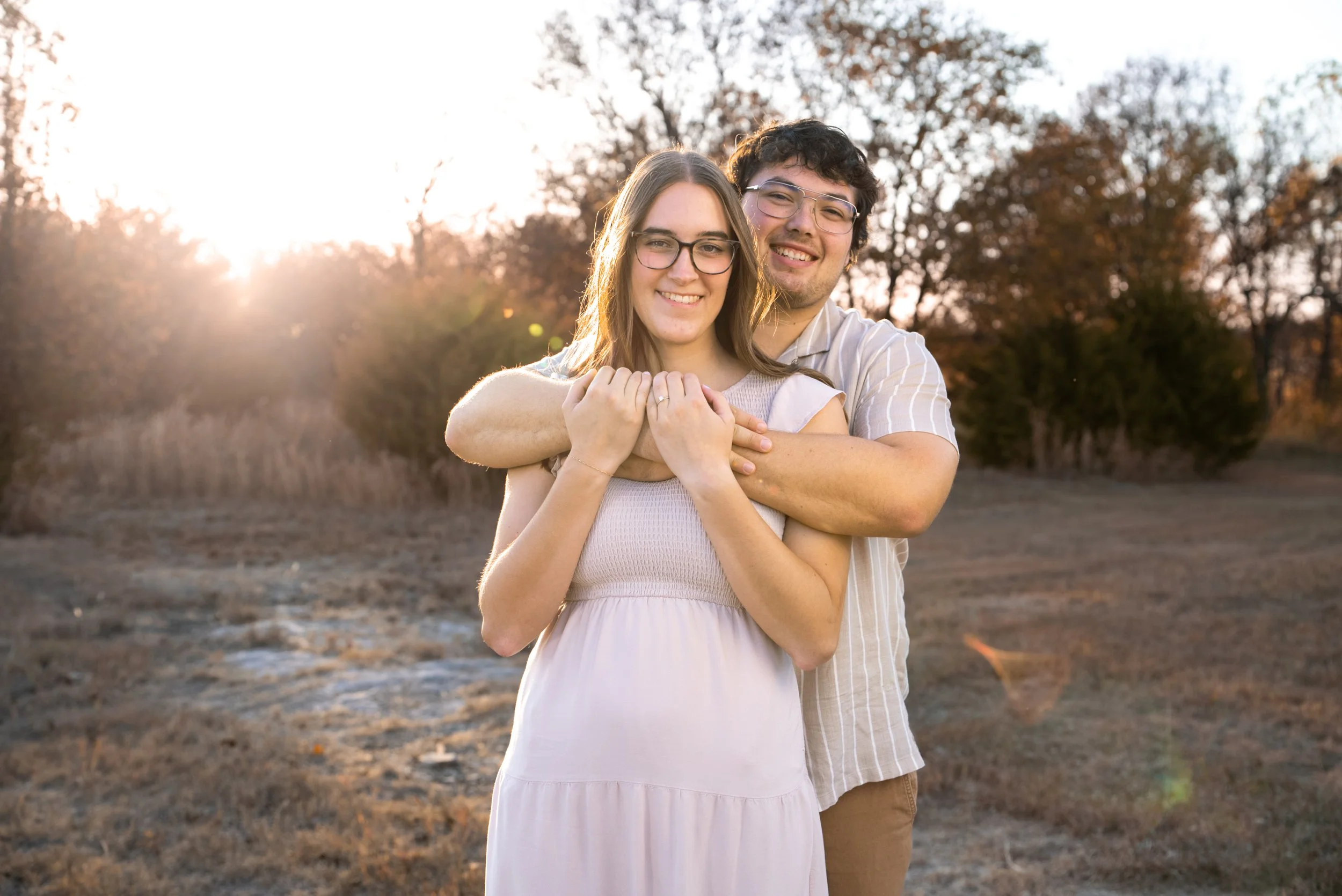 A young couple standing outdoors during sunset, embracing and smiling at the camera. The woman is wearing a light-colored dress and the man is wearing glasses and a short-sleeved shirt.