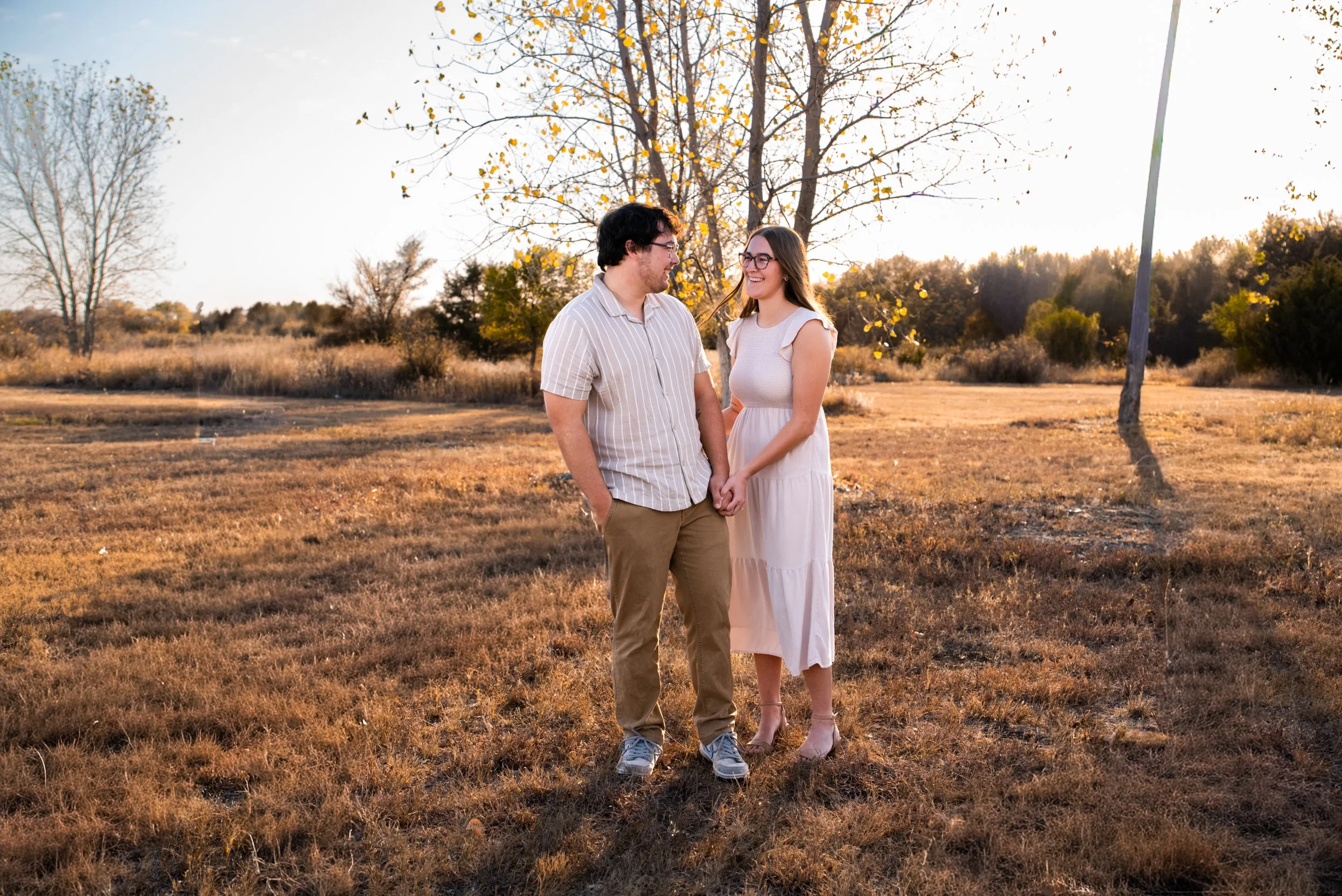 A couple holding hands and smiling at each other outdoors on a sunny autumn day, surrounded by dry grass and trees with yellow leaves in the background.
