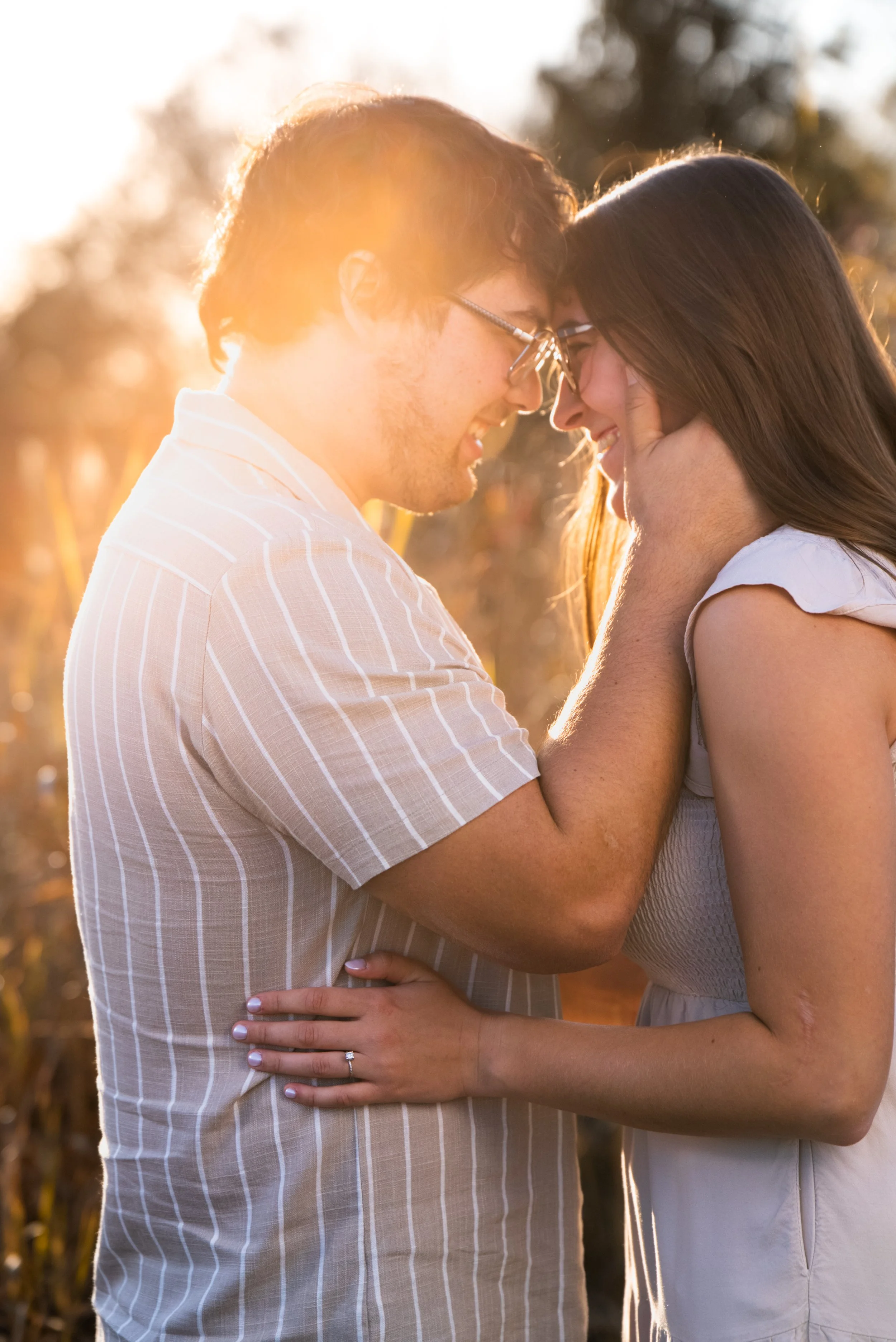 A couple smiling and touching foreheads outdoors during sunset, with sunlight glowing behind them and blurred trees in the background.