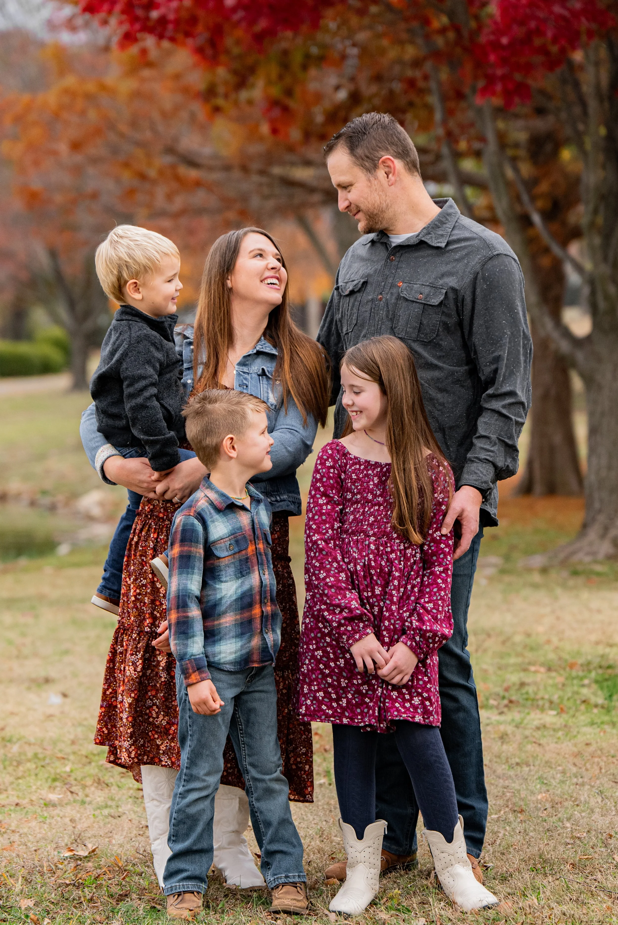 Family of six outdoors during fall, standing on grass with trees with autumn leaves in background. The family includes two adults and four children. The woman and girl are smiling at each other, while the man and two boys are looking at the woman. Th