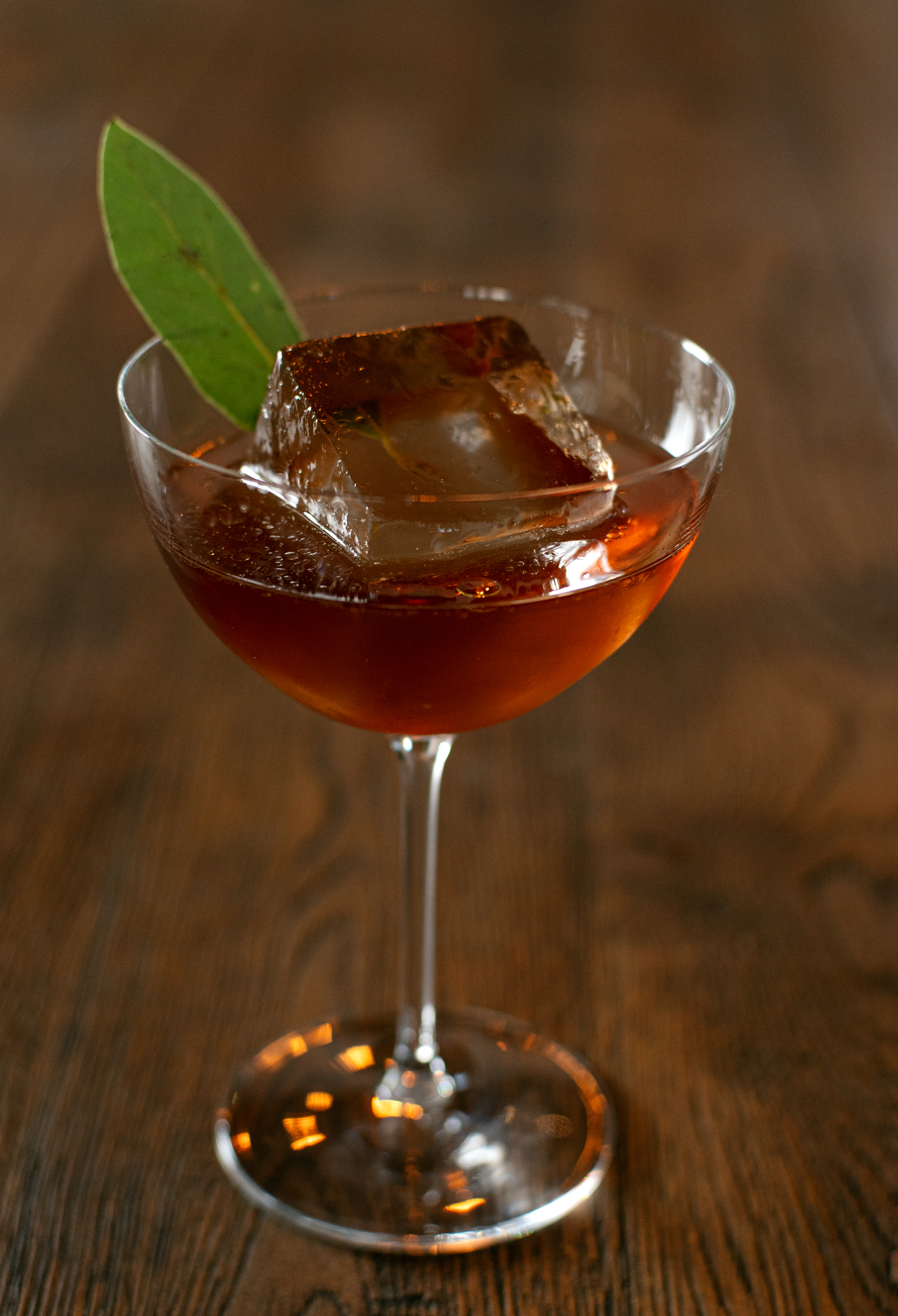 Refined cocktail served in a coupe glass with a large clear ice cube and a green leaf garnish, presented on a wooden surface in a warm, elegant setting.