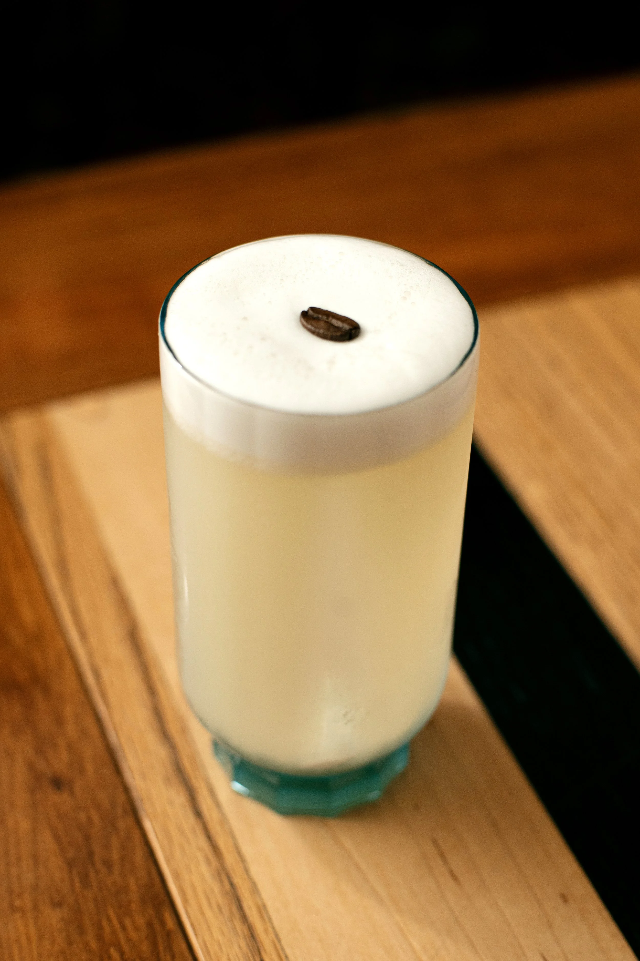 Signature cocktail with a creamy white foam, garnished with a single coffee bean, served in a minimalist glass on a wooden bar surface.