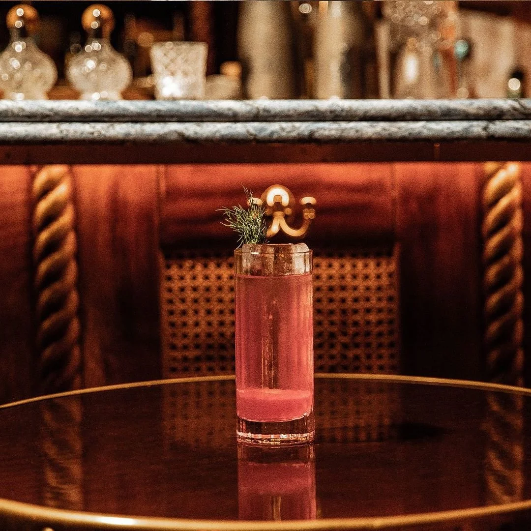 A pink cocktail in a tall glass garnished with greenery and a decorative stir stick on a reflective dark surface, with a background of wooden furniture and decorative glass bottles.