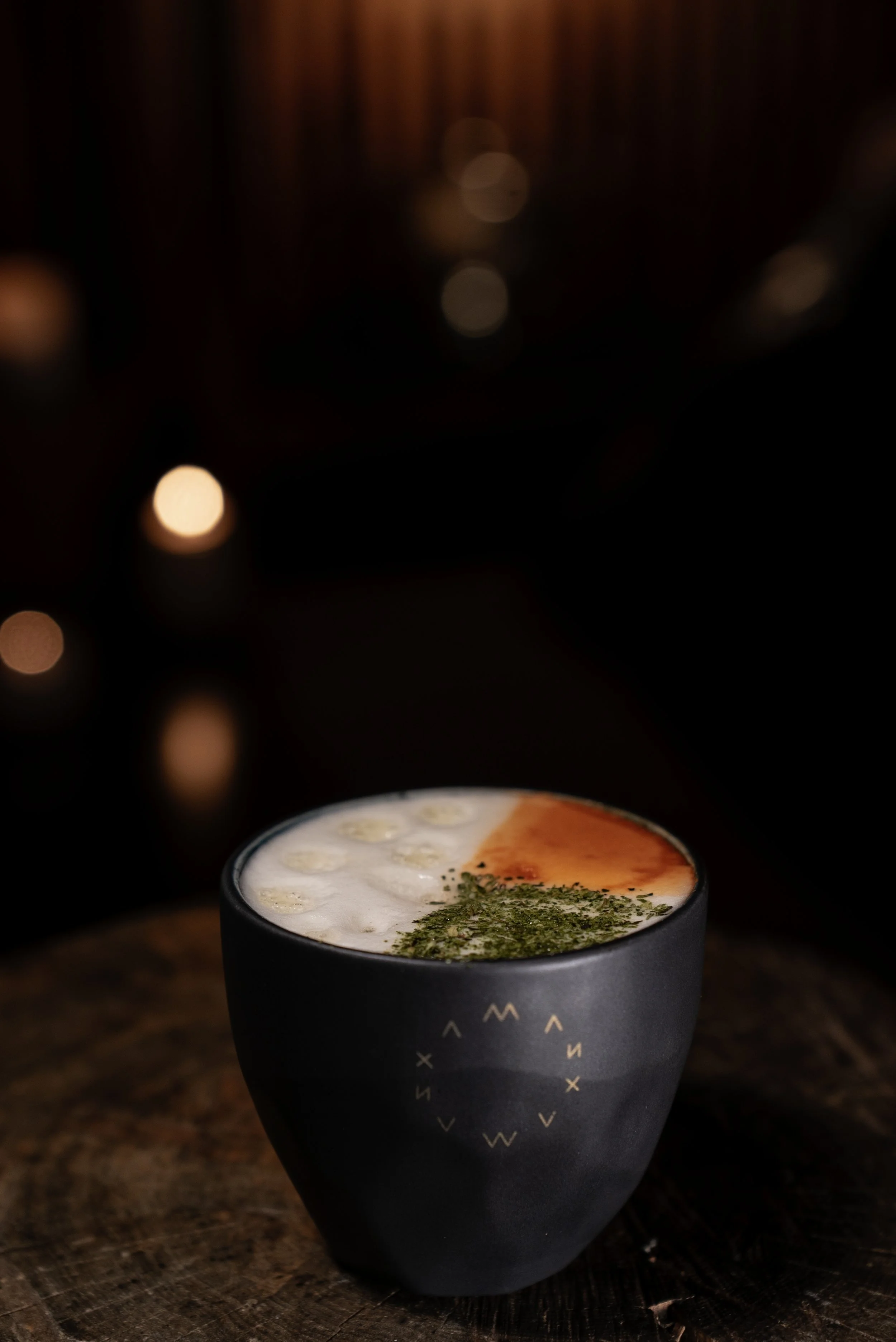 Elegant signature cocktail served in a matte black ceramic cup, topped with creamy foam, citrus accents, and fresh green herbs, photographed in a warm, intimate bar setting.