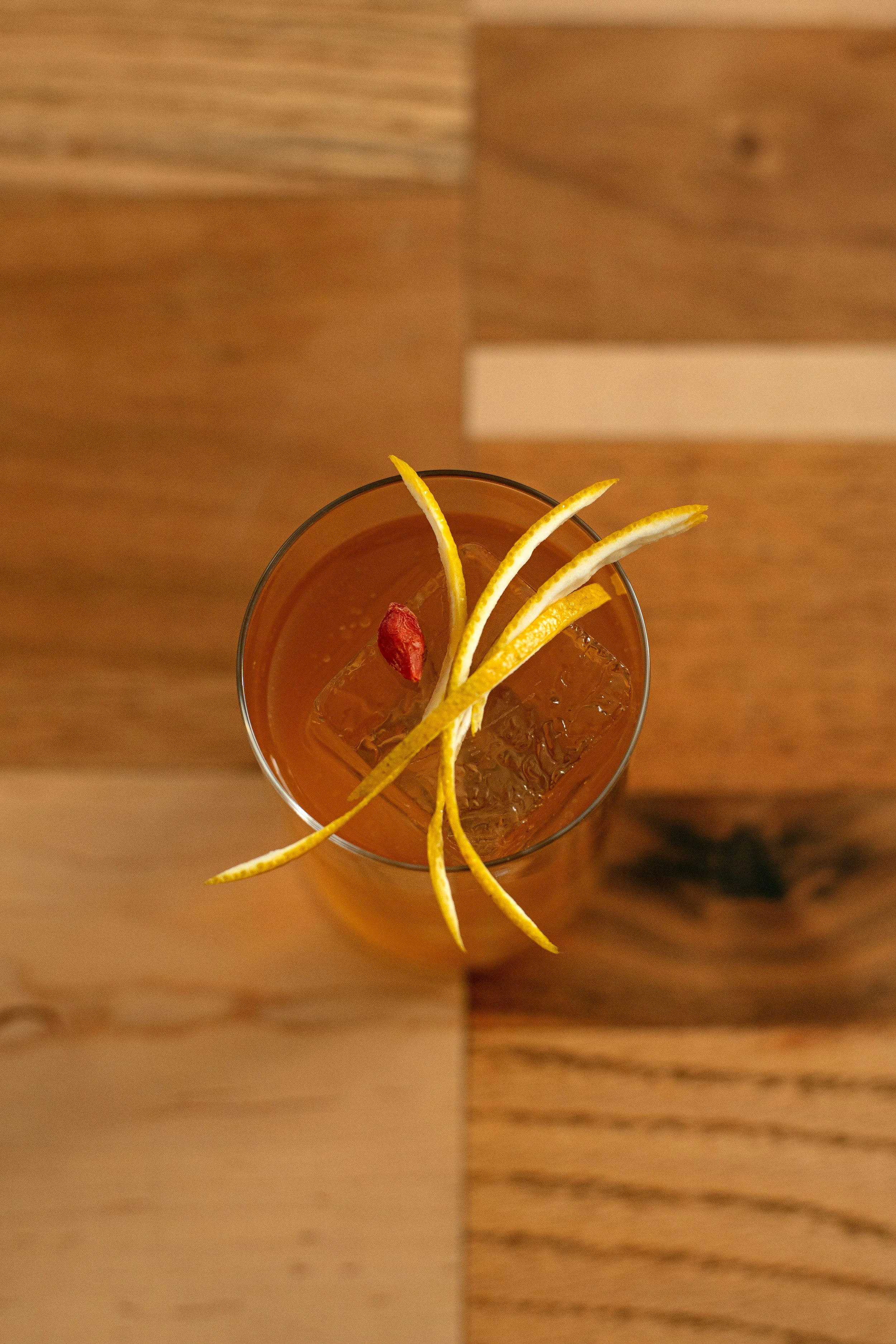 Top-down view of a cocktail in a glass with lemon twists and a red berry, served over ice, on a wooden table.