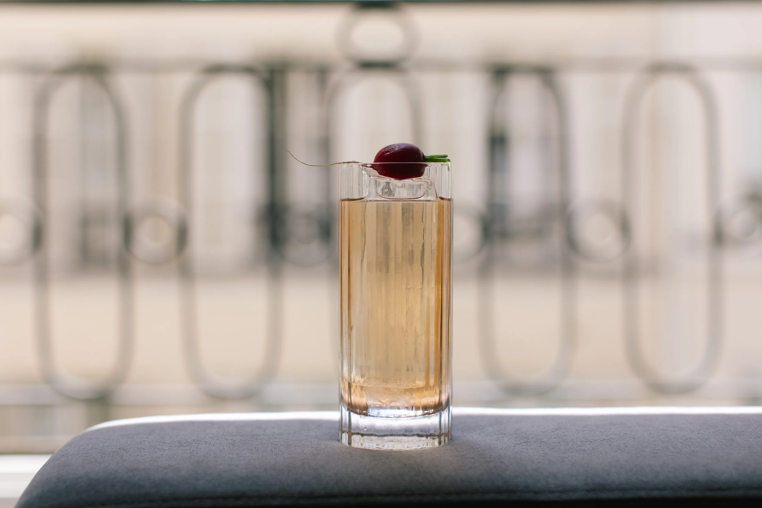 A tall, clear glass filled with a light amber liquid, topped with a cherry and a small green garnish, placed on a dark surface with a blurred background of a metallic structure.