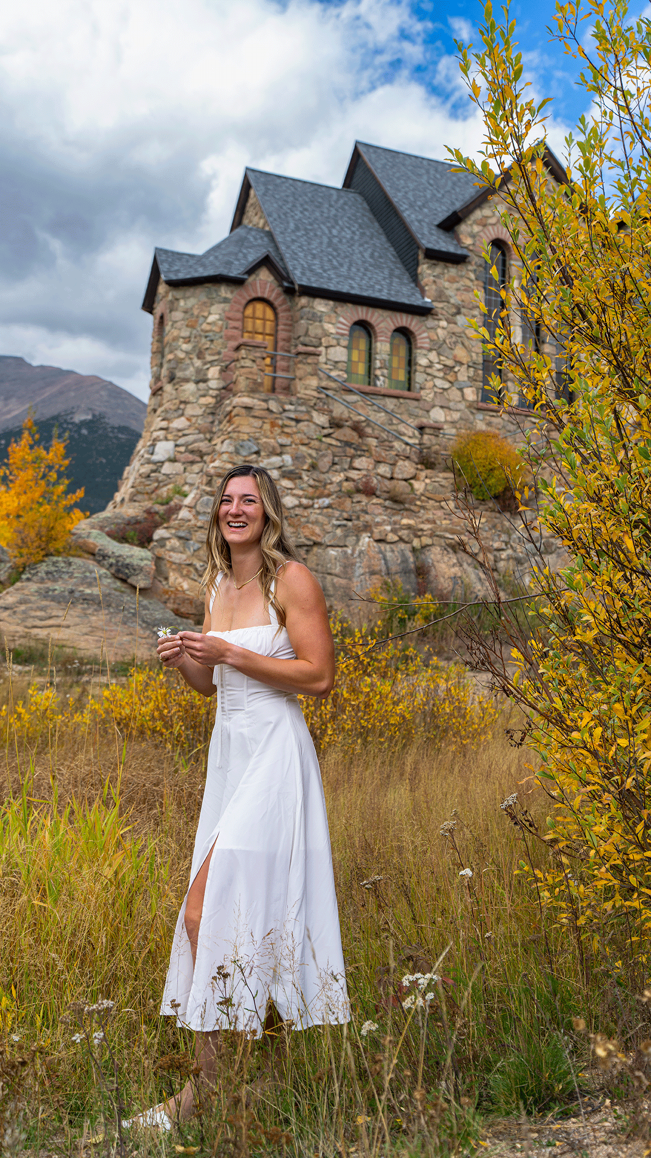 A woman in a white dress smiling and holding a small flower, standing in a field of tall grass and yellow bushes in front of a stone house with a steep roof, in a mountainous landscape with a cloudy sky.