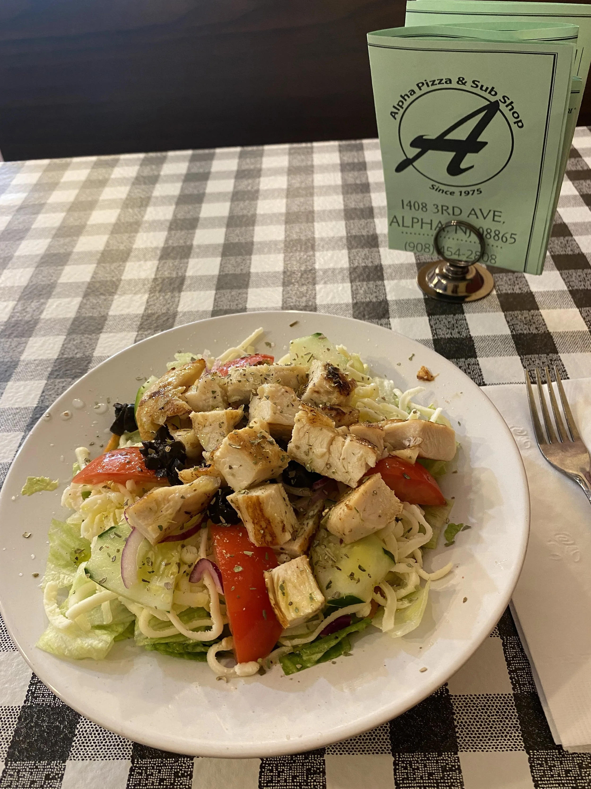 A plate of salad with grilled chicken, lettuce, tomato, cucumber, and red onion, on a checkered tablecloth, with a fork, napkin, and green paper menu in the background.