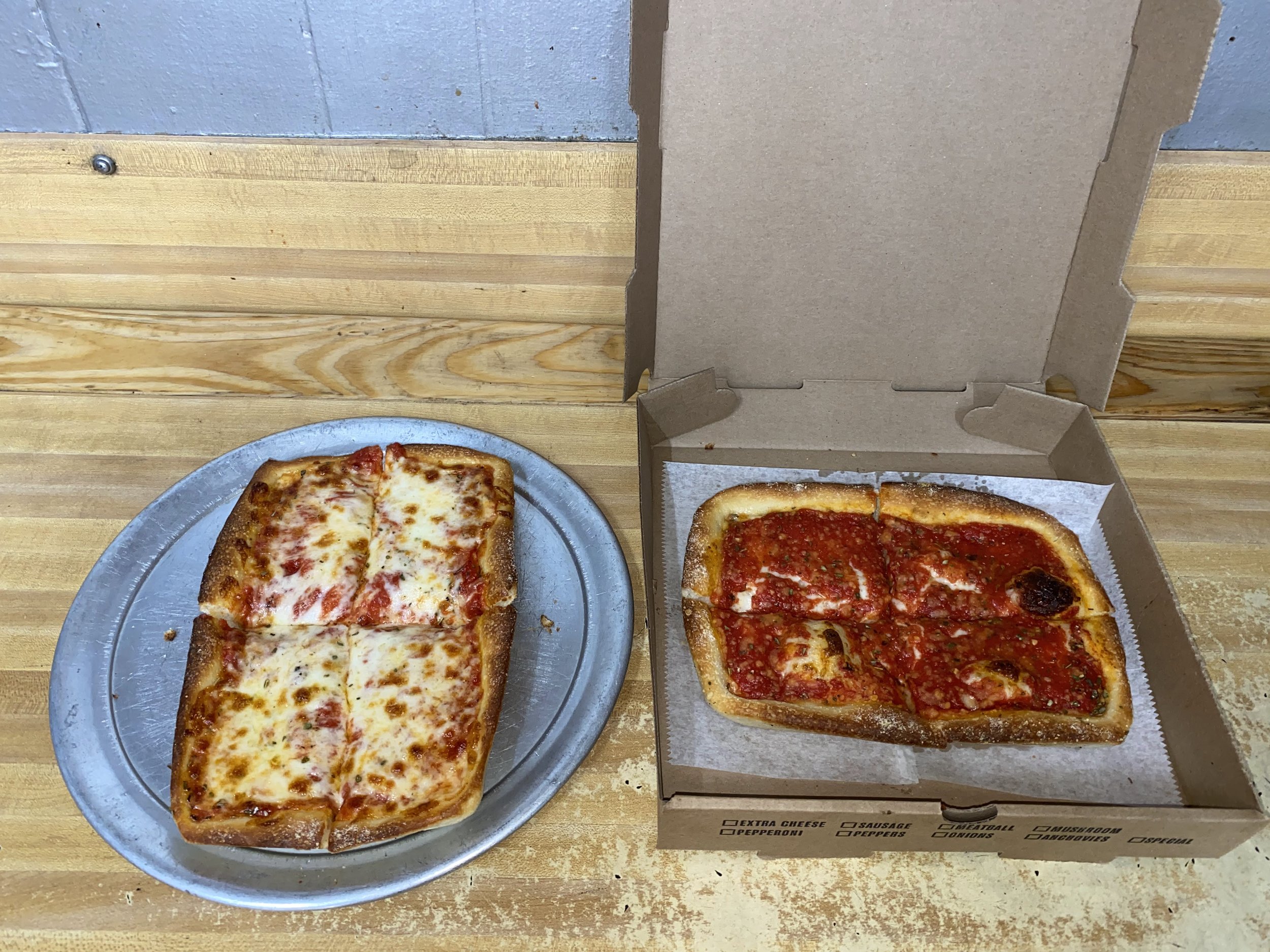 Two slices of cheese pizza on a metal tray, and another two slices of cheese pizza in a cardboard pizza box on a wooden table.