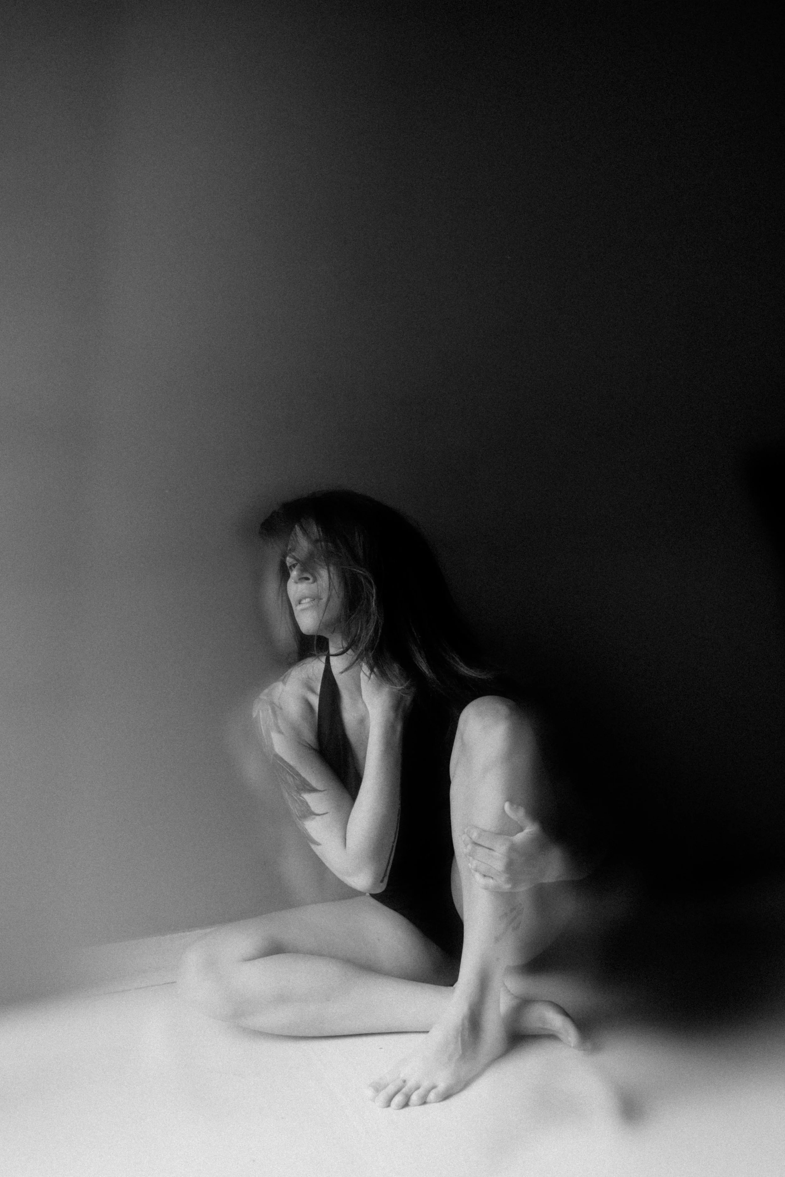 A black-and-white photo of a woman sitting on the floor, with her arms wrapped around her legs and her head turned to the side, looking to the left. She appears to be topless.