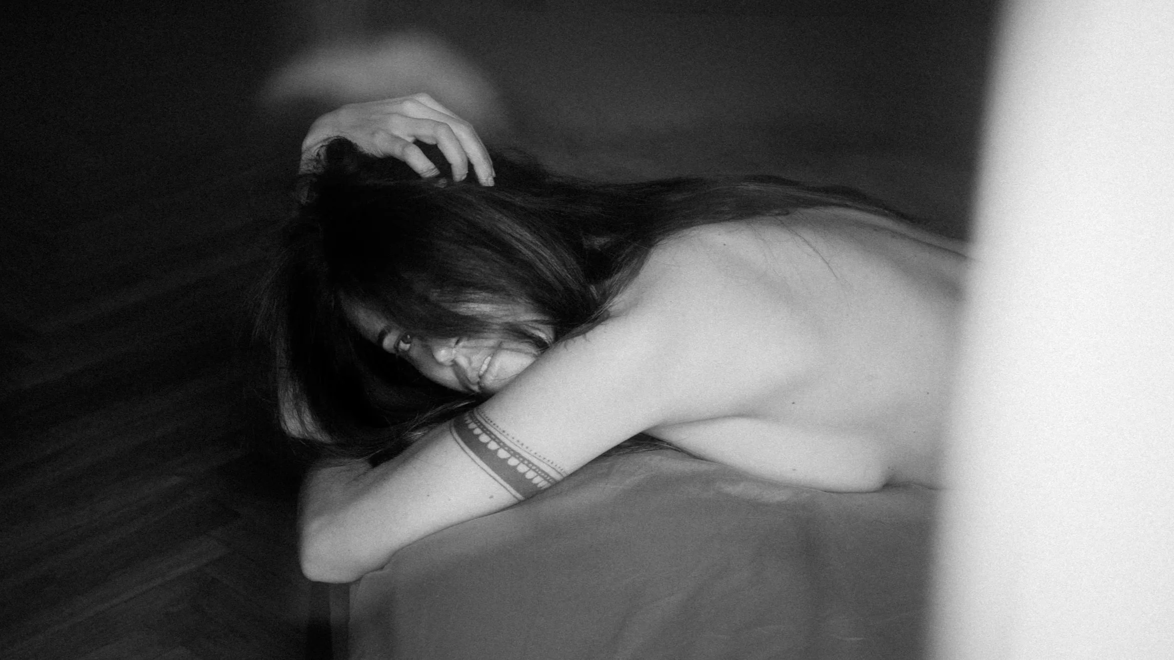 A person with long dark hair lying on the floor, smiling with eyes closed, hand on head, wearing a sleeveless top and arm bracelet, in a black-and-white photo