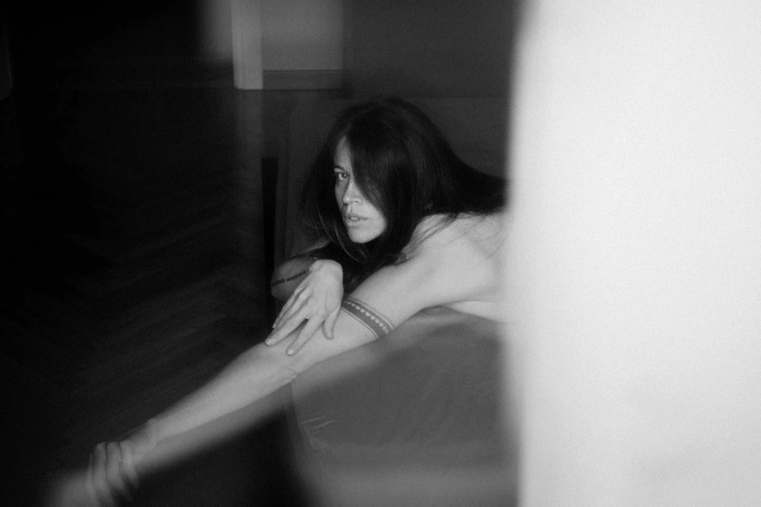 A black and white photo of a woman with long dark hair, lying on the floor, partially obscured by shadows and a white wall, looking directly at the camera.