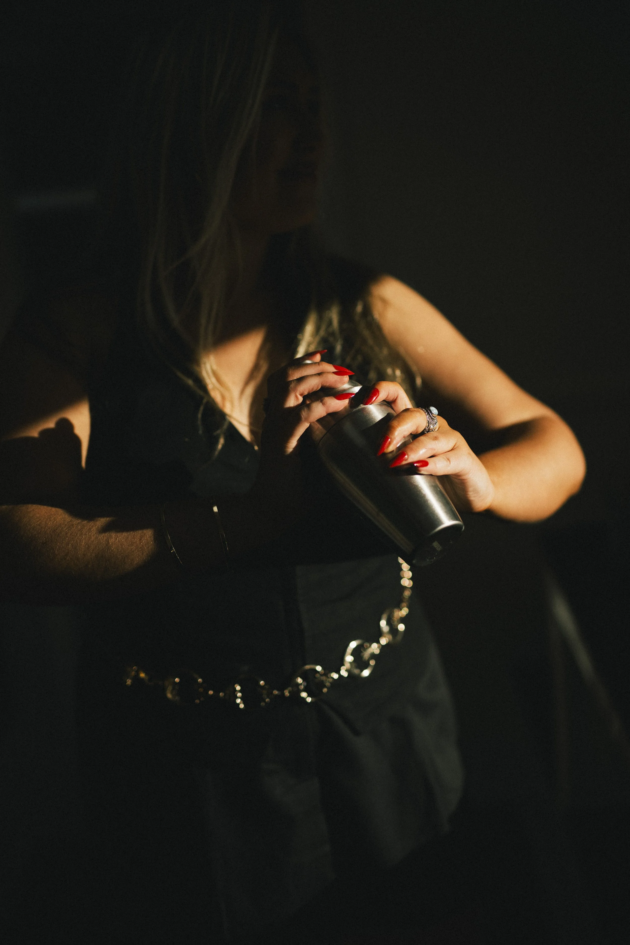 A woman holding a metallic tumbler with a straw in a dark setting, illuminated by soft light highlighting her hands with red nail polish, jewelry, and part of her outfit.