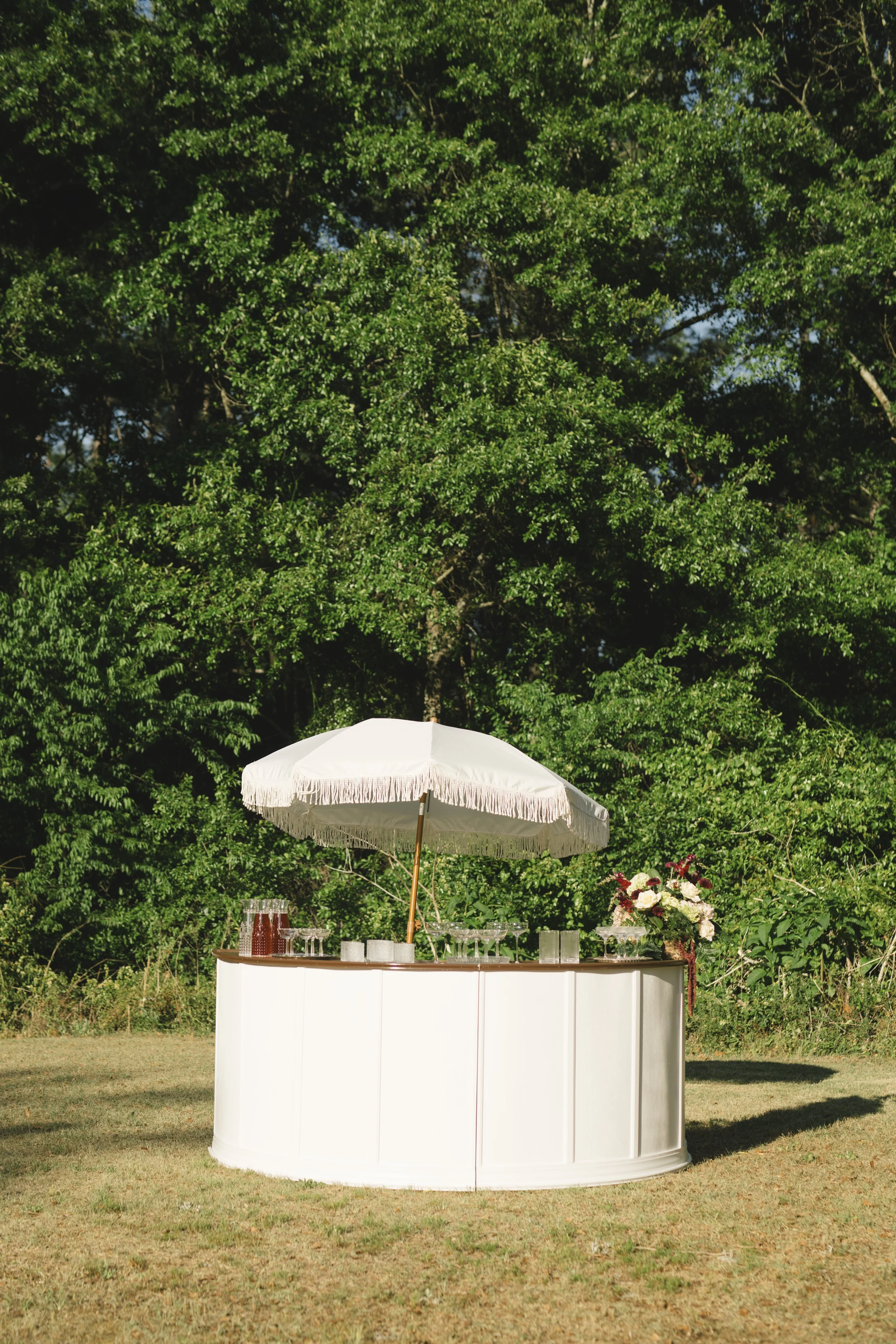A white circular bar setup with an umbrella and glassware on it, set outdoors on a grassy area with green trees in the background.