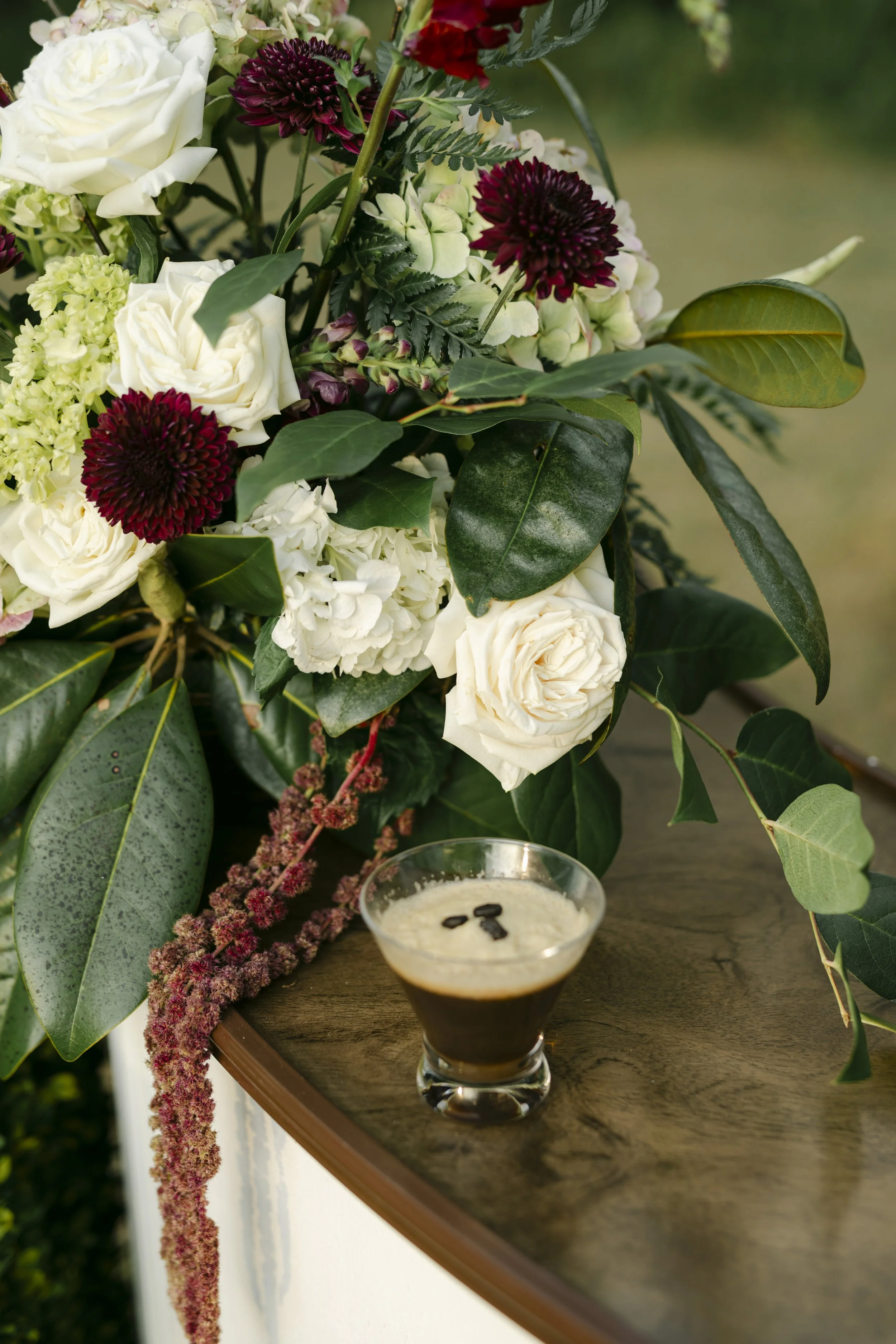 A floral arrangement with white roses, maroon dahlias, and green leaves on a wooden table, with a glass of coffee with whipped cream and coffee beans on top in the foreground.