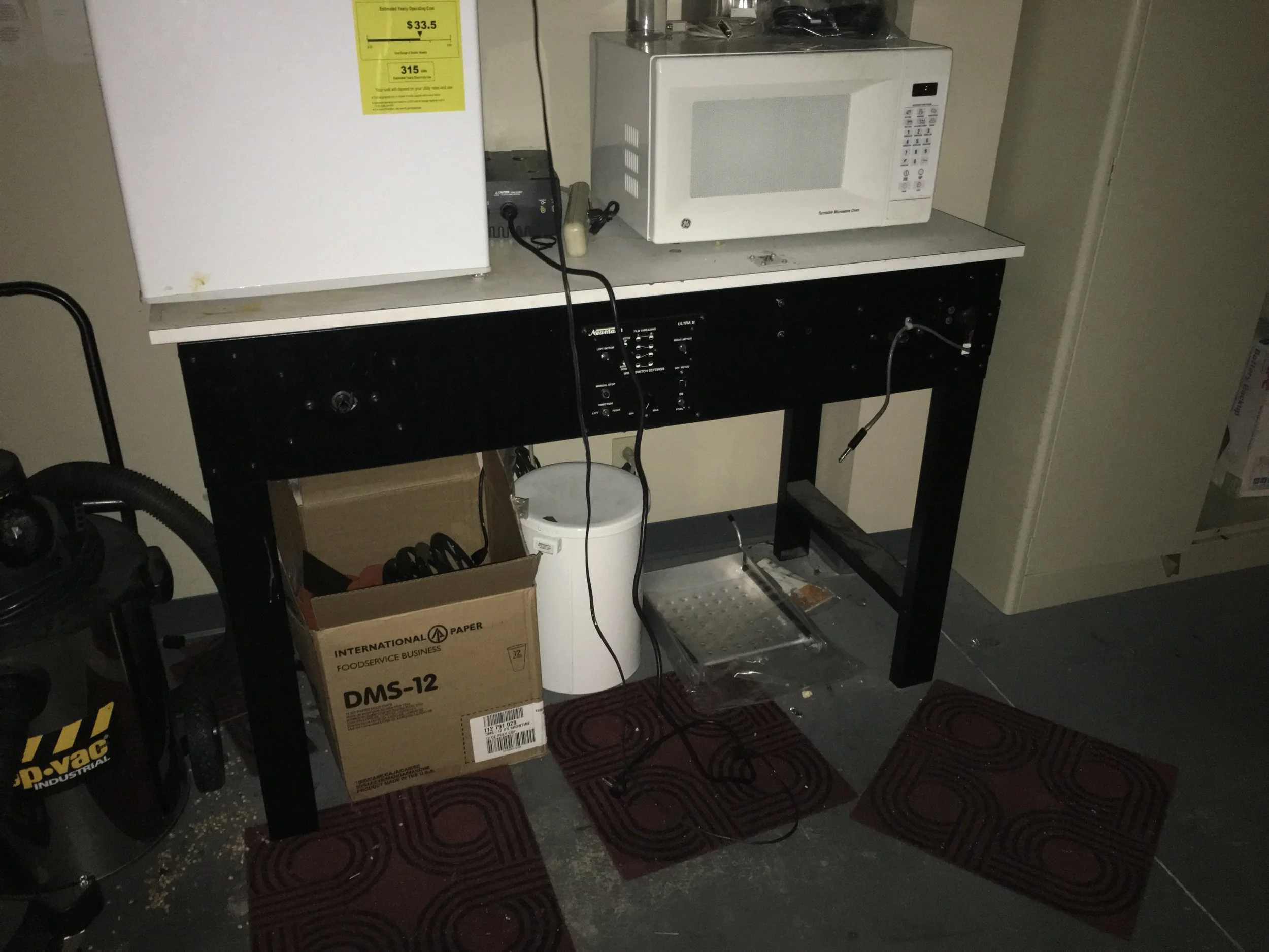 A workspace with a black workbench, a white microwave on top, an open box of supplies, a white trash bin, and red patterned floor mats on a concrete floor.