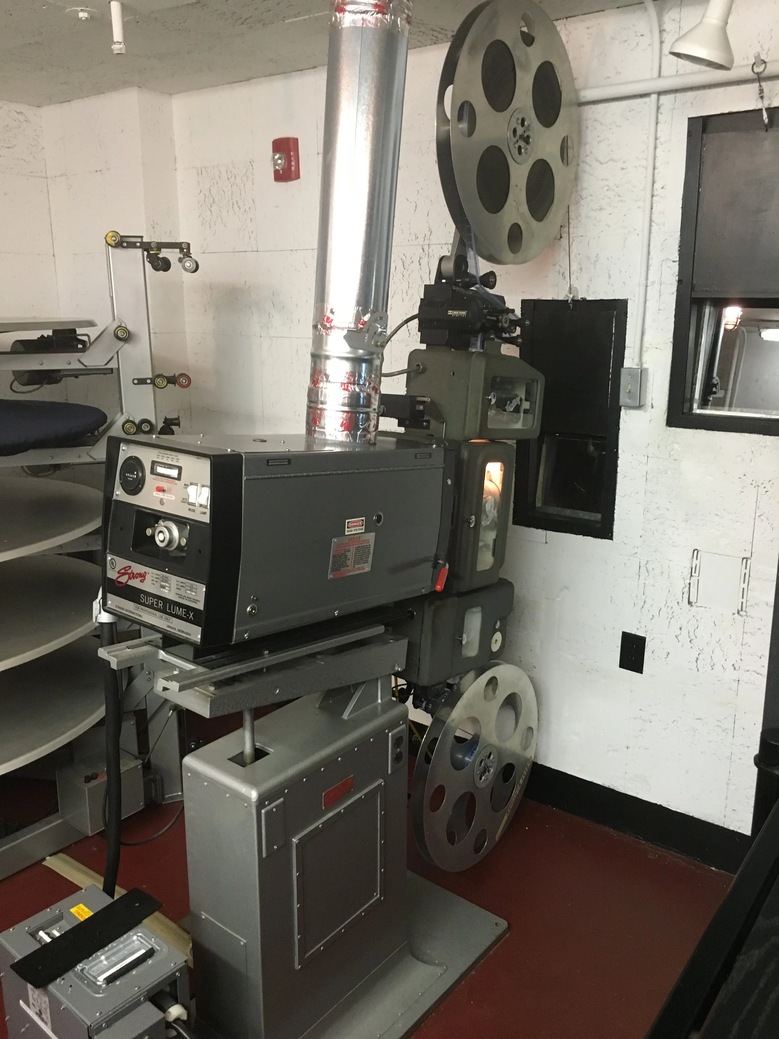 A vintage film projector with large reels, positioned on a stand, in a room with white textured walls and a red floor.