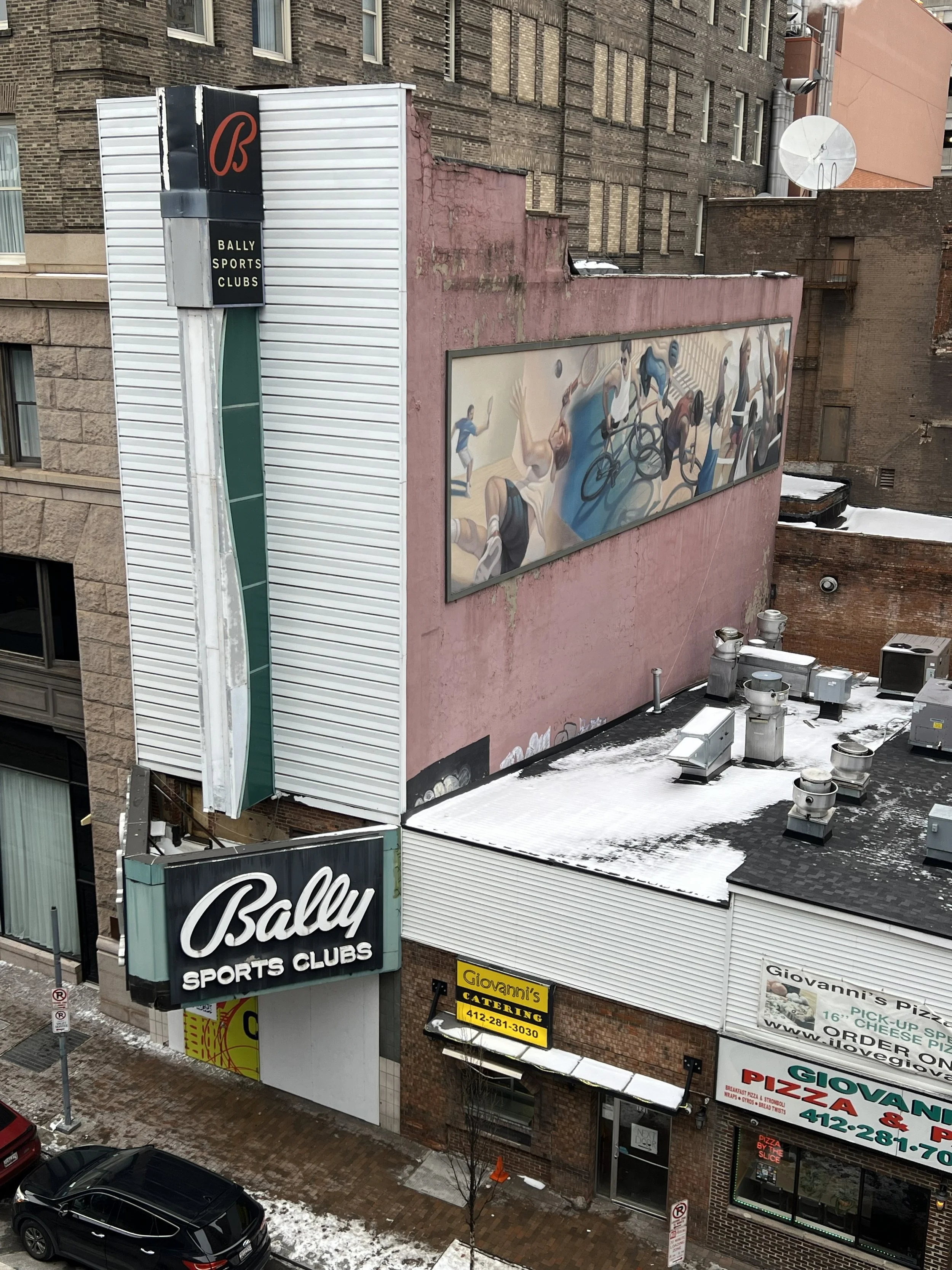 An urban scene showing the exterior of Bally Sports Clubs with a large sign and mural, building rooftops with snow, parked cars, and various business signs including Giovanni's Catering and Pizza.