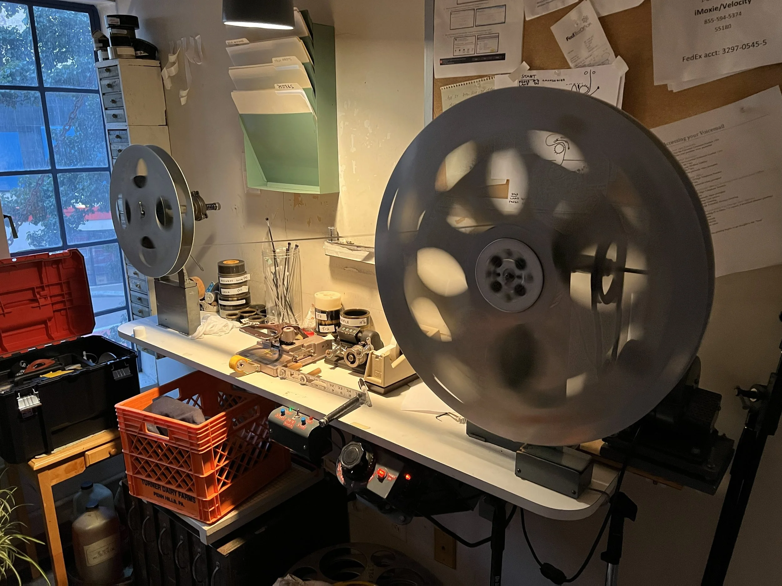 A workspace with film reels, film projectors, and various equipment on a white table, next to a window with a view of trees outside.