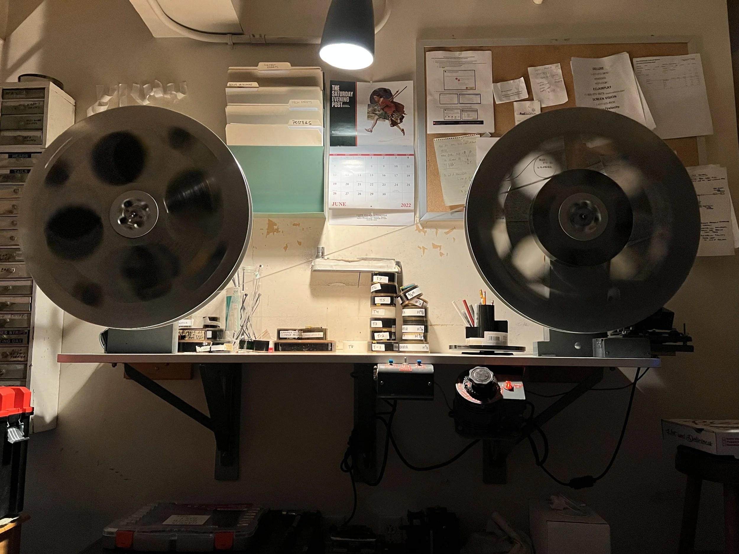 Image of a workspace with a large reel-to-reel tape recorder on a table. The table also has small boxes, cables, and various office papers. A wall behind the table has a calendar, a corkboard with notes, and a wall-mounted lamp illuminating the scene