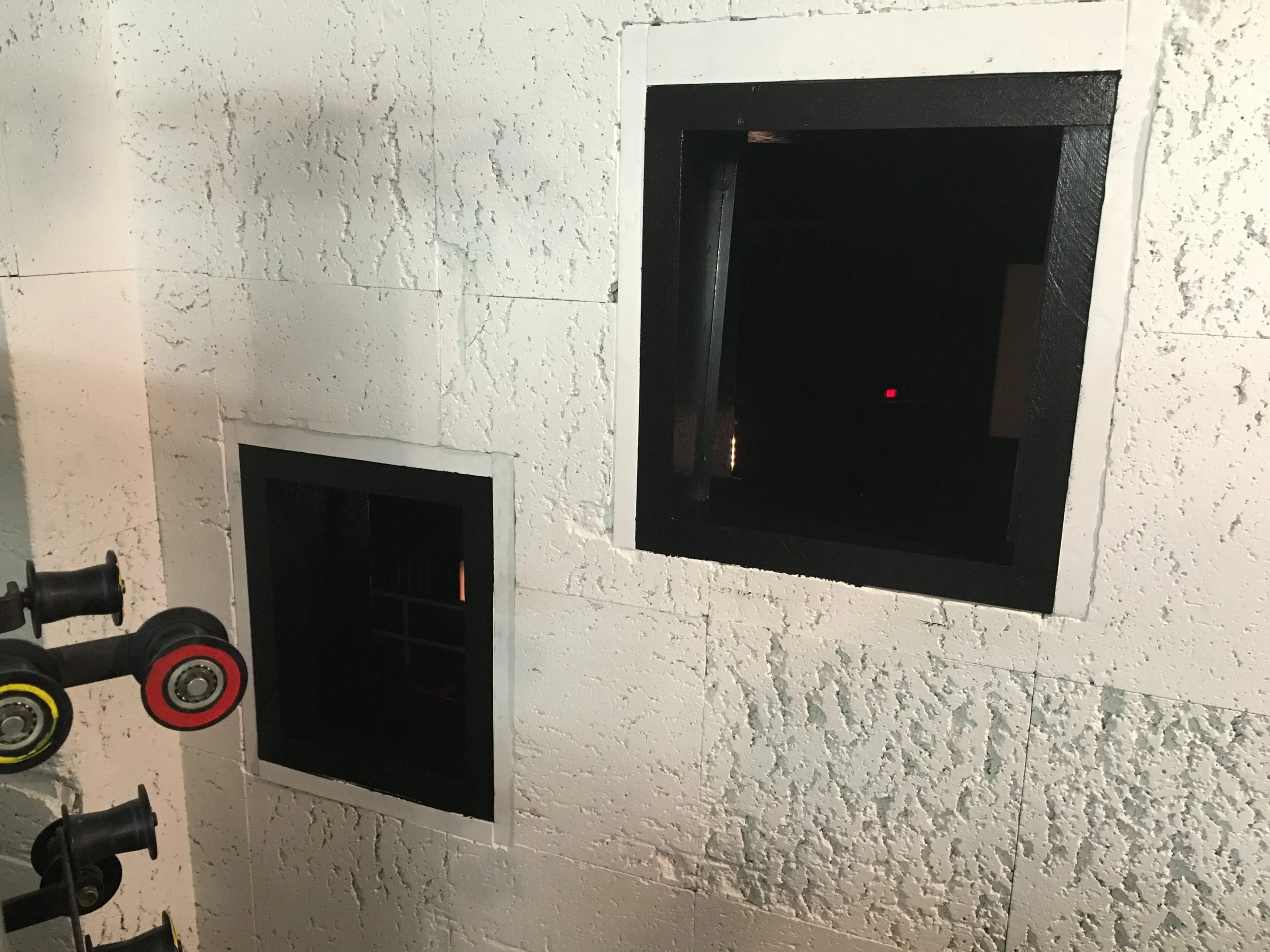 A wall with two black framed openings, one smaller and one larger, and some tools with black and red handles hanging on the left.