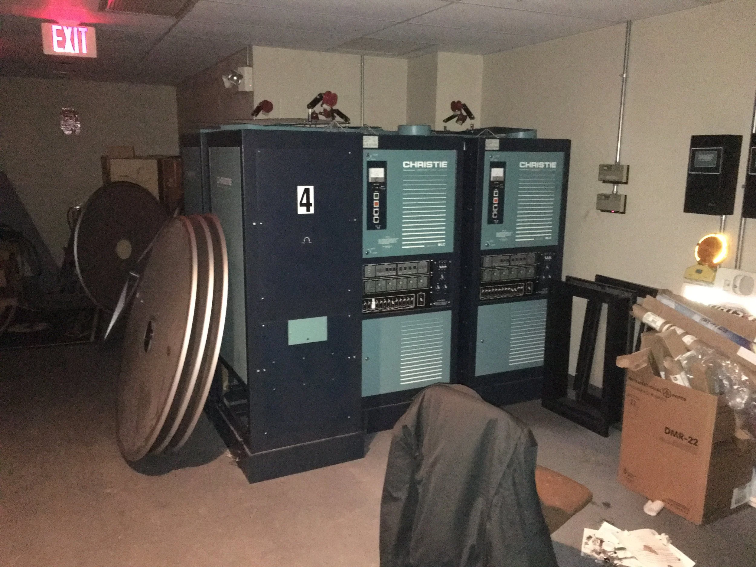Two large blue electrical control panels labeled 'CHRISTIE' are against a wall in a cluttered room. There are large reels of cable leaning against the panels and a cardboard box filled with various items on the floor nearby. An illuminated red 'EXIT'