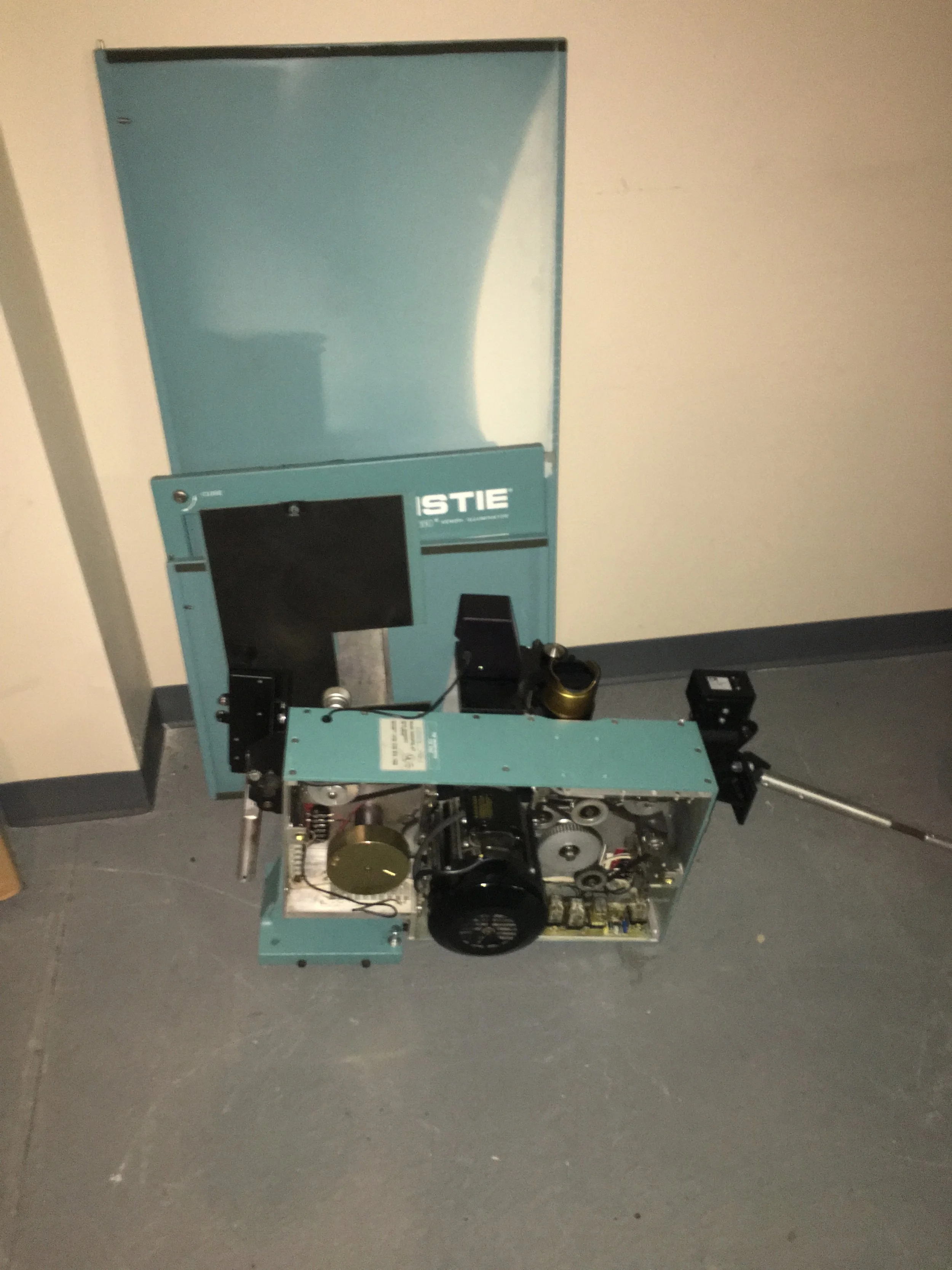 Disassembled machine with exposed internal parts and a blue cover, placed on a gray floor against a light-colored wall.