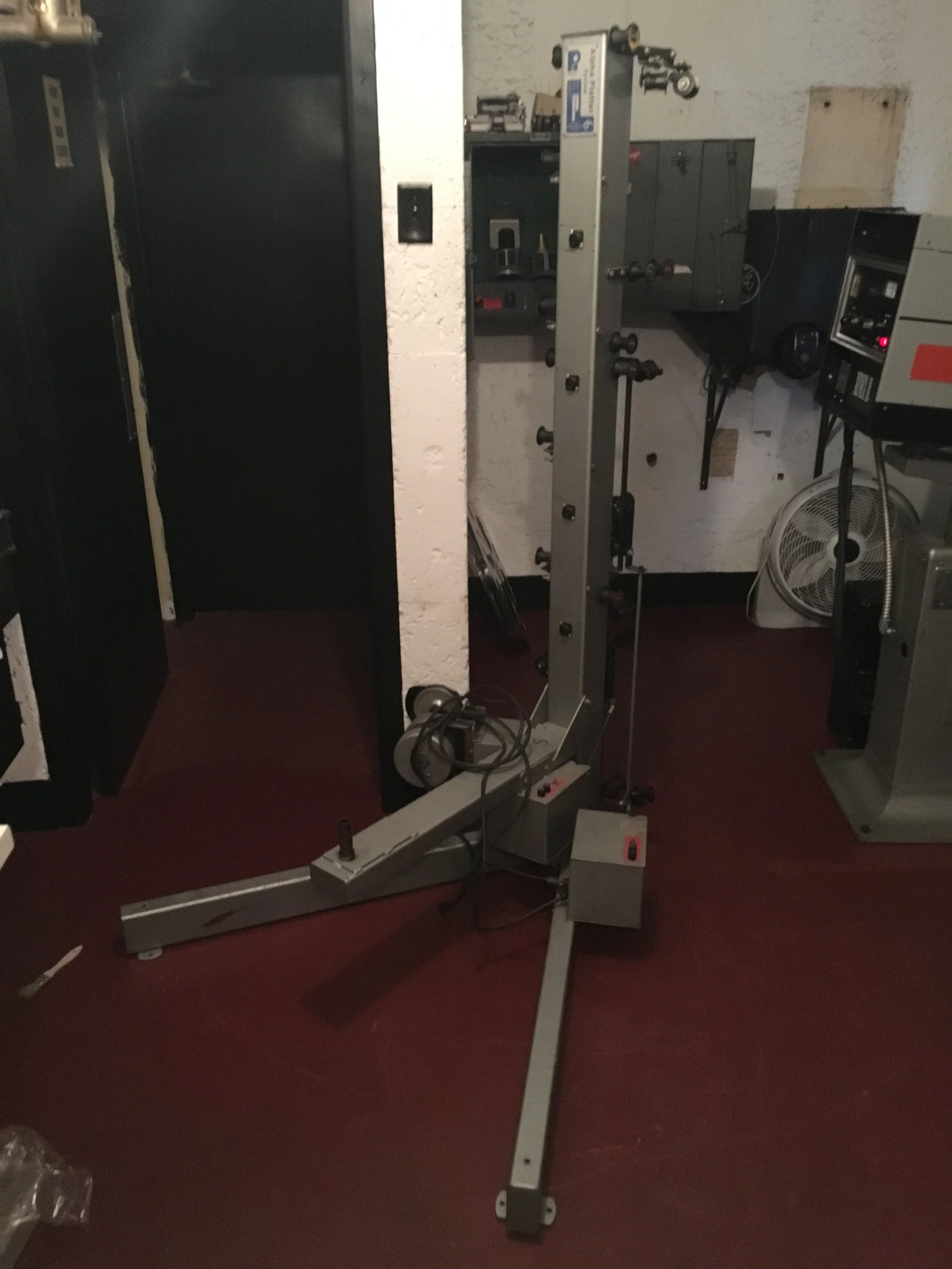 Metal device with wheels and adjustable arms, possibly a piece of industrial or scientific equipment, situated in a room with various electronic devices and a fan in the background.