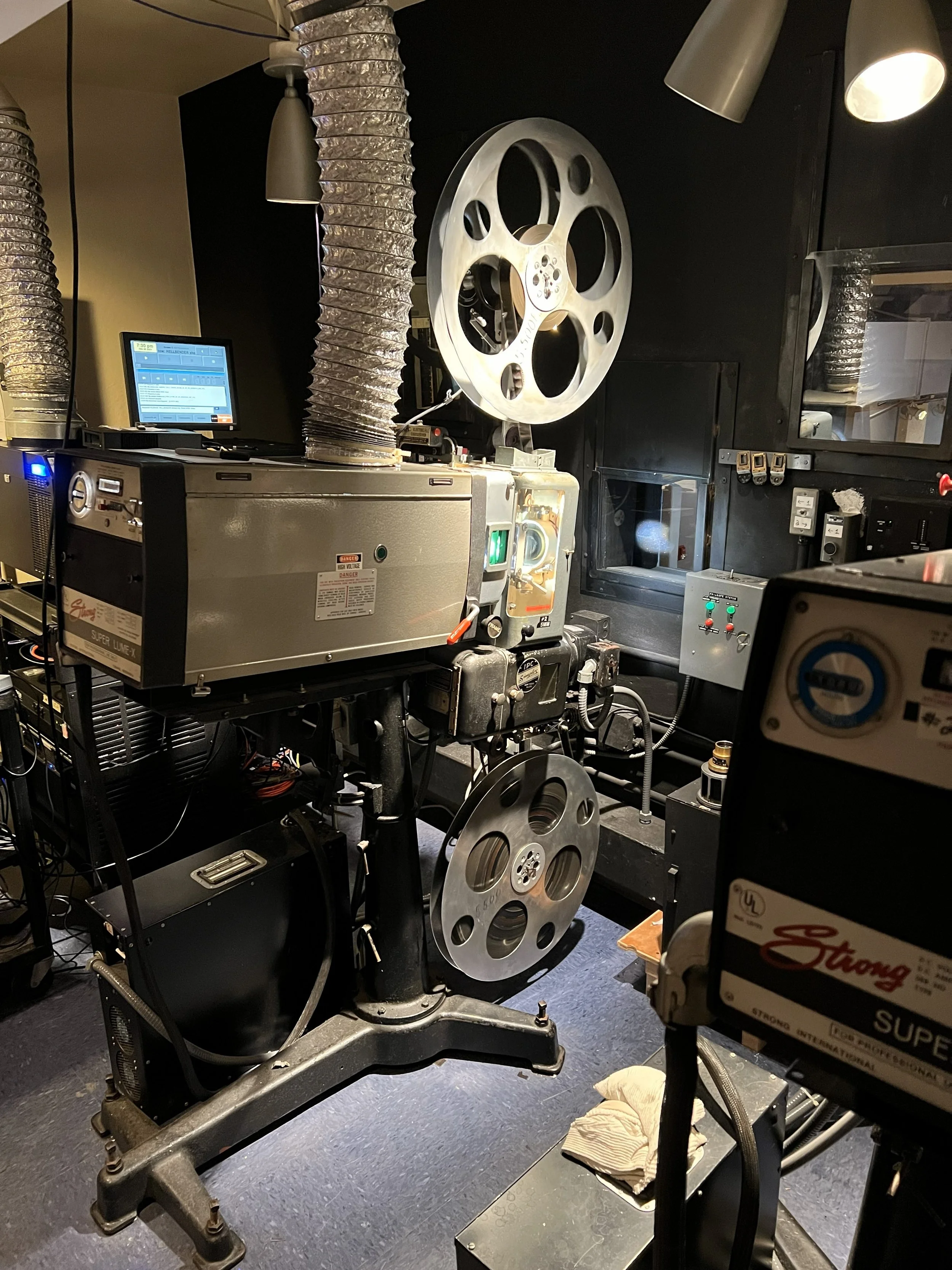 A film projection setup with two large metal reels, film projector, and ventilation tubes, in a dimly lit room.
